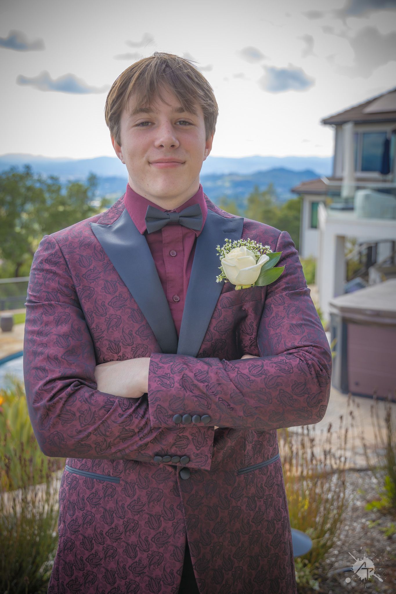 Person in burgundy patterned suit with boutonniere poses against mountain scenery.
