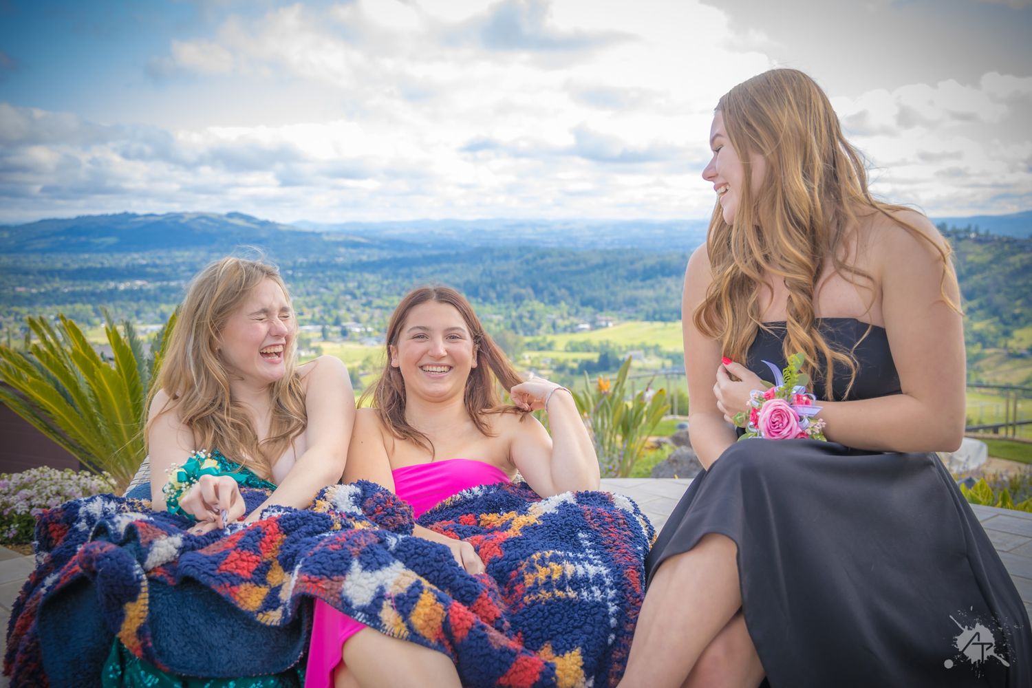 Three people in formal dresses share a candid moment on mountain terrace.