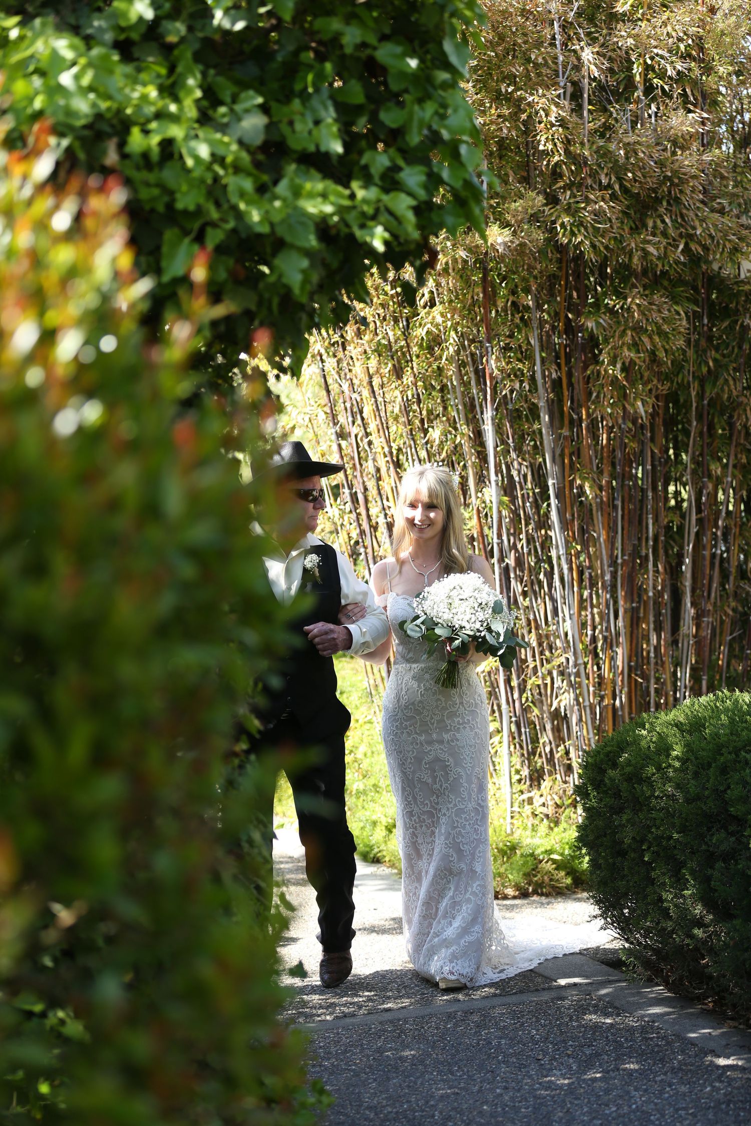 Bride and escort walk along a garden path with bamboo and greenery in the background on a sunny day.