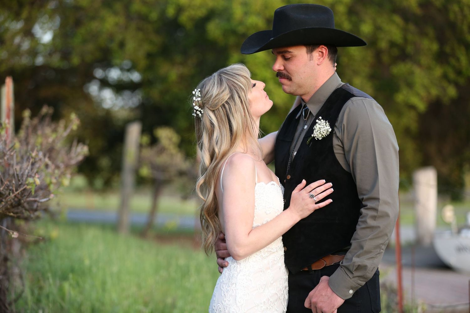 A romantic western-themed wedding couple embraces outdoors surrounded by green foliage.
