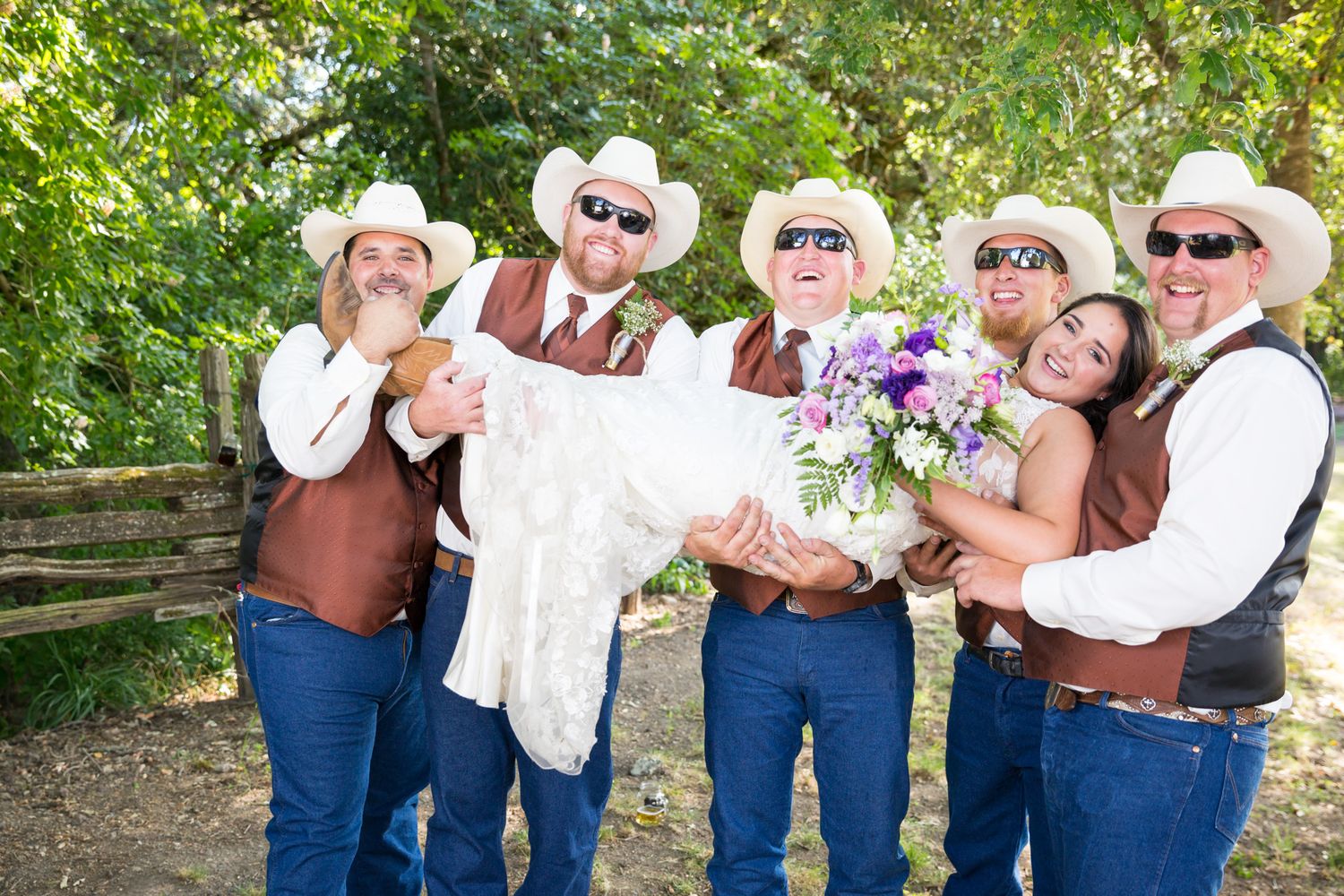 A festive wedding group wearing brown vests and cowboy hats poses playfully with a wedding dress outdoors.