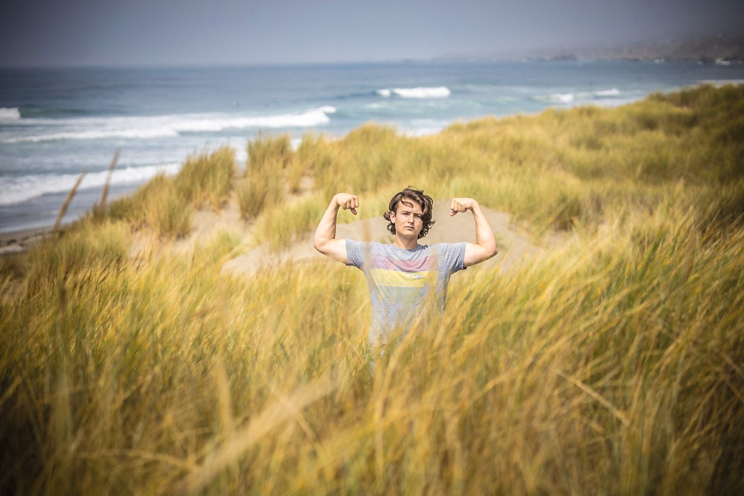 Figure with raised arms joyfully standing among coastal dunes with ocean waves behind.