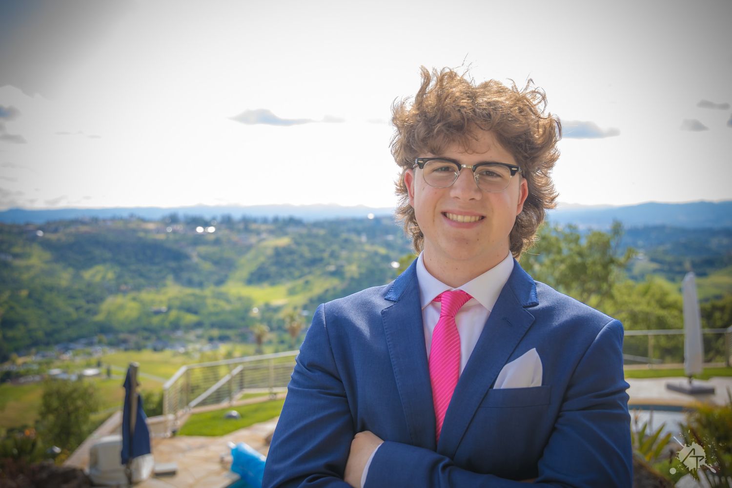 Person wearing blue suit with pink tie poses confidently against mountain landscape.