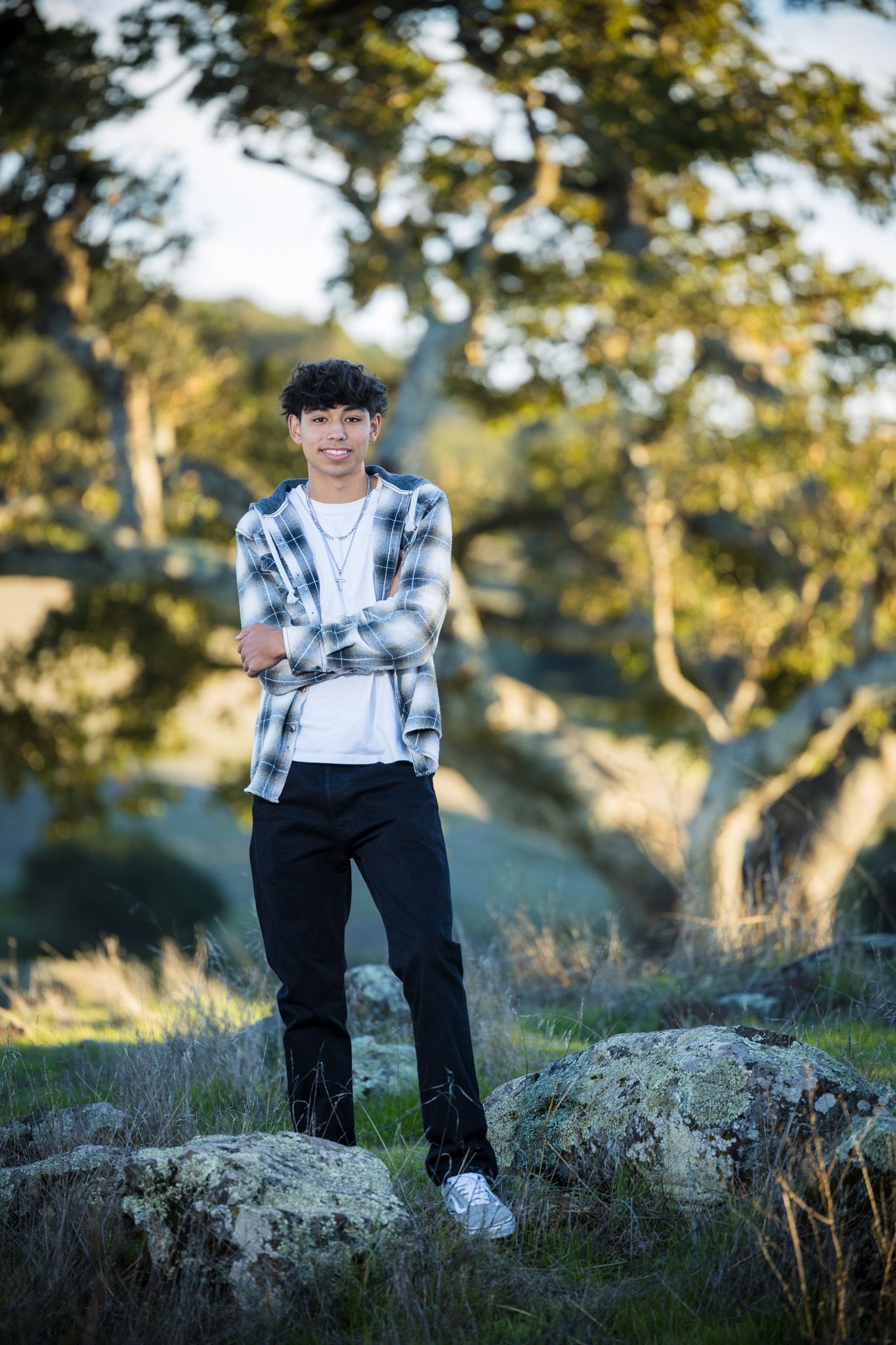 Senior Photos - Person in plaid shirt and dark pants sitting casually on a large rock during golden hour in nature.