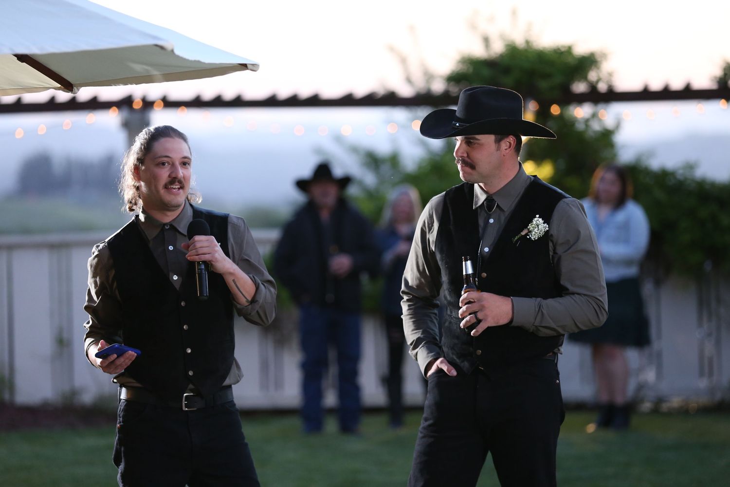 Two people in dark western attire dance together outdoors at dusk while others watch in the background.