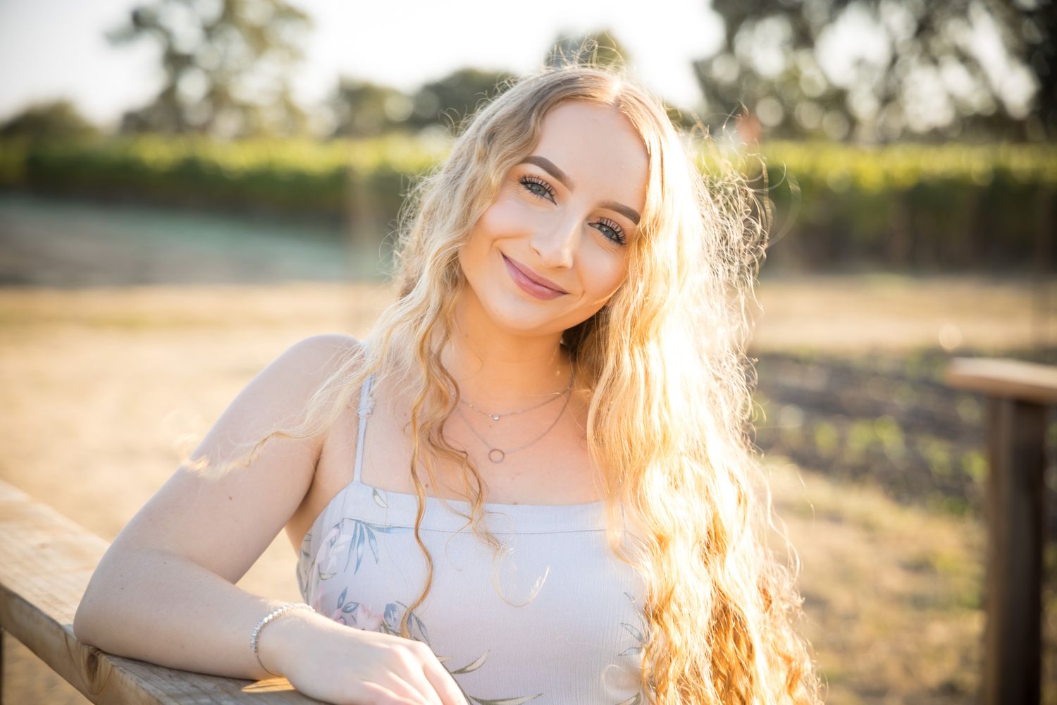 Backlit portrait of a person with long blonde hair in a white dress leaning against a wooden fence at sunset.