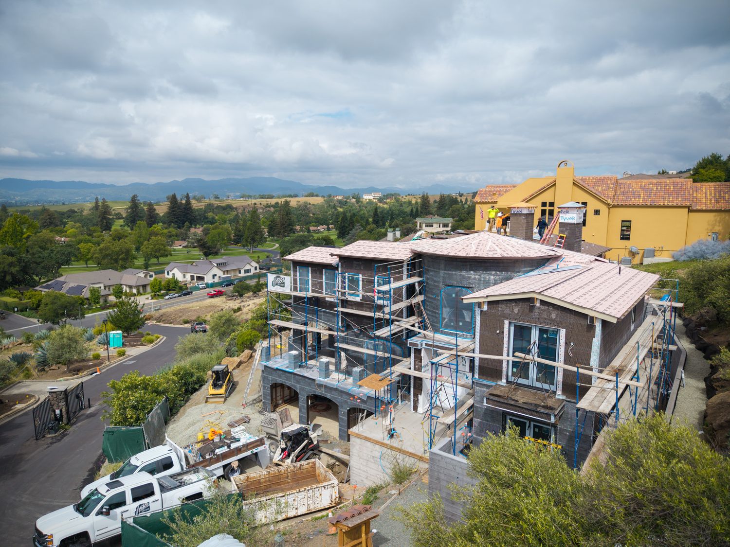 Aerial view of a multi-story home under construction with scaffolding, surrounded by trees and hills under cloudy skies.