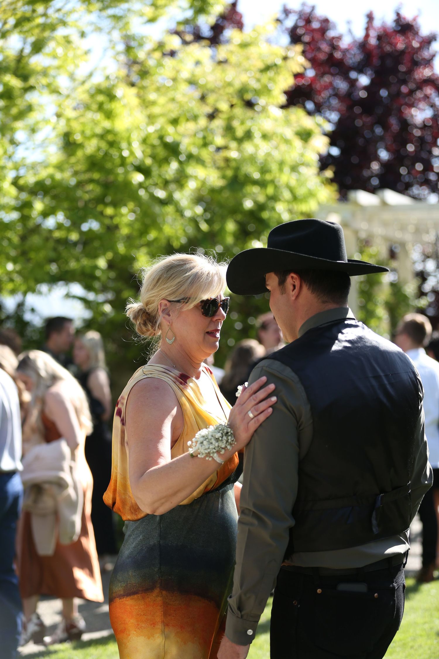 Two people interact outdoors at a summer gathering with green trees and other attendees visible in the background.