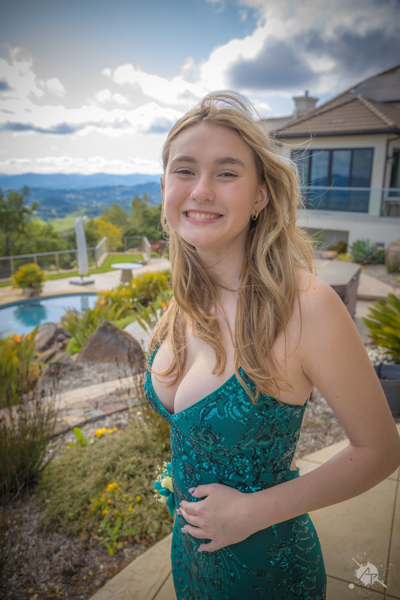 Person in teal lace formal dress poses outdoors with mountain backdrop and swimming pool.