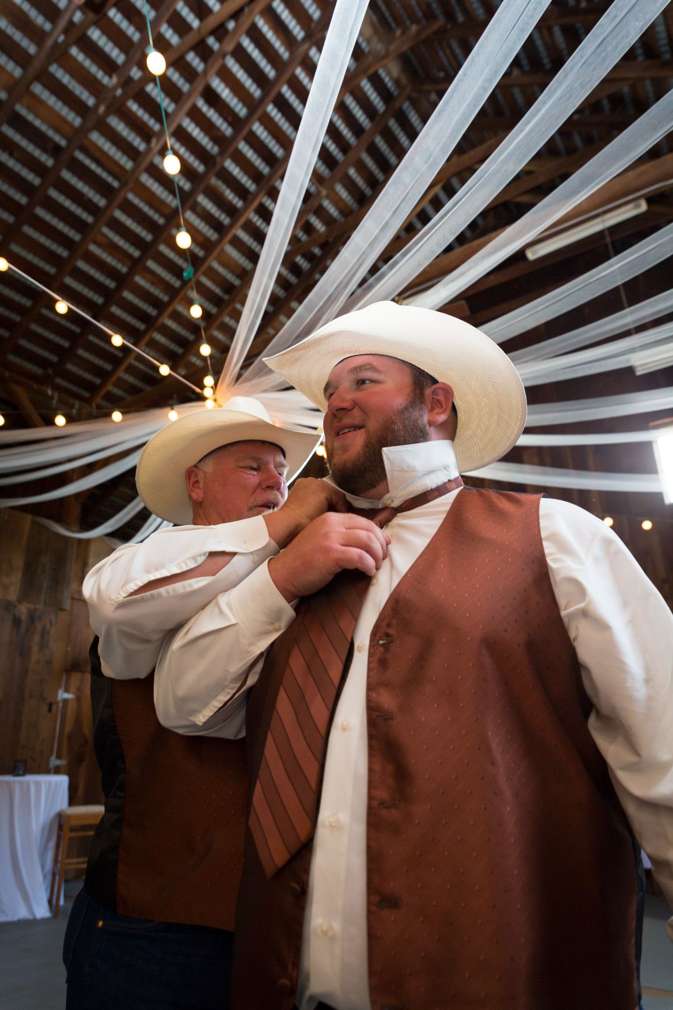 Two men in cowboy hats and western attire share a lighthearted moment adjusting a tie inside a decorated barn venue.