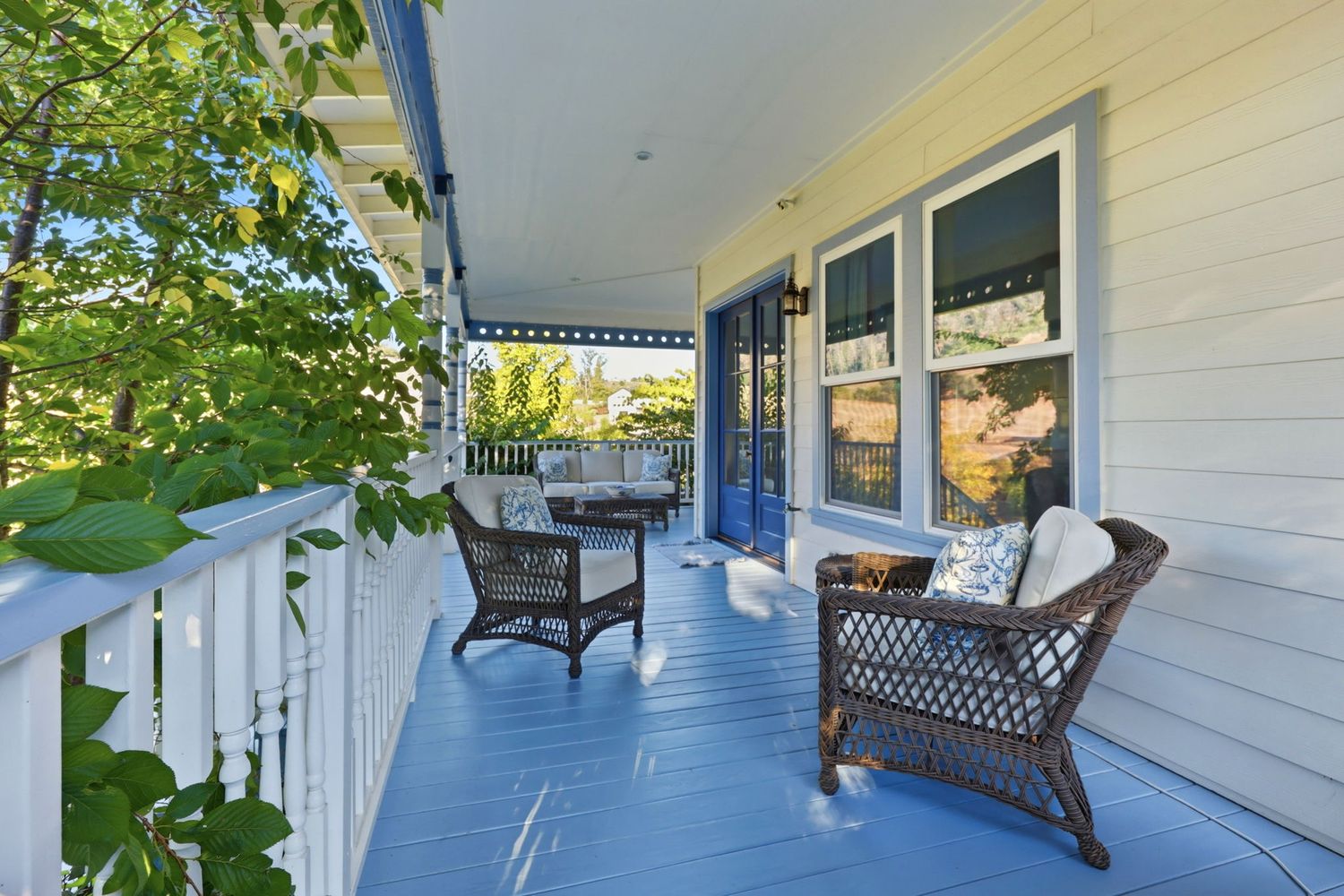 Charming blue-painted front porch with wicker chairs, white railings, and lush green trees surrounding the home.