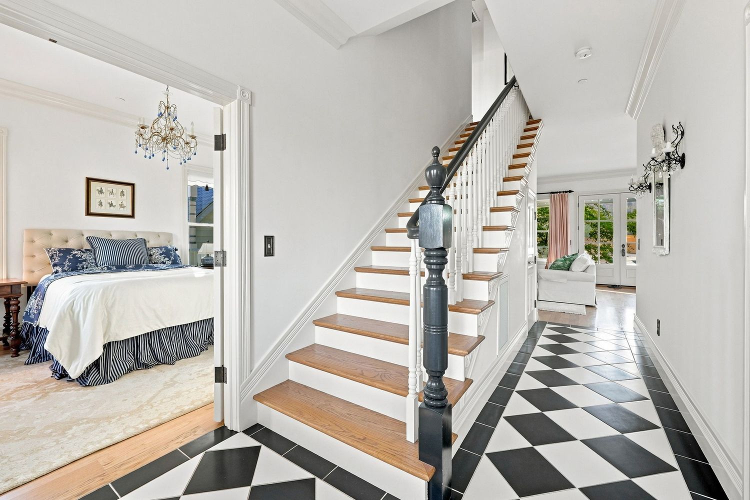 Elegant foyer with black and white checkered floors, a grand staircase, chandelier, and a glimpse into a blue bedroom.