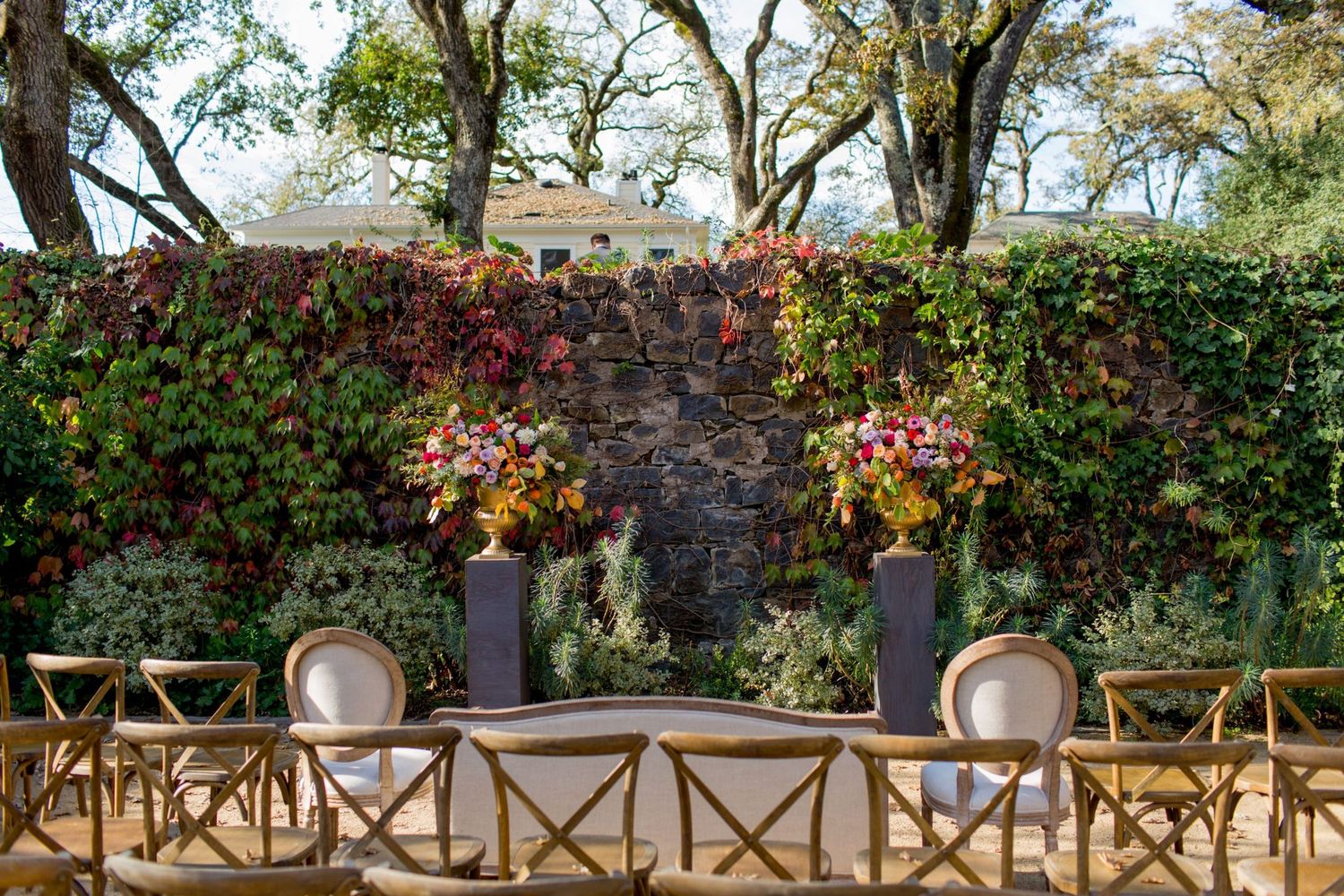 A rustic outdoor patio with wooden pergola, stone walls, and vibrant red flowering plants creating a cozy seating area.