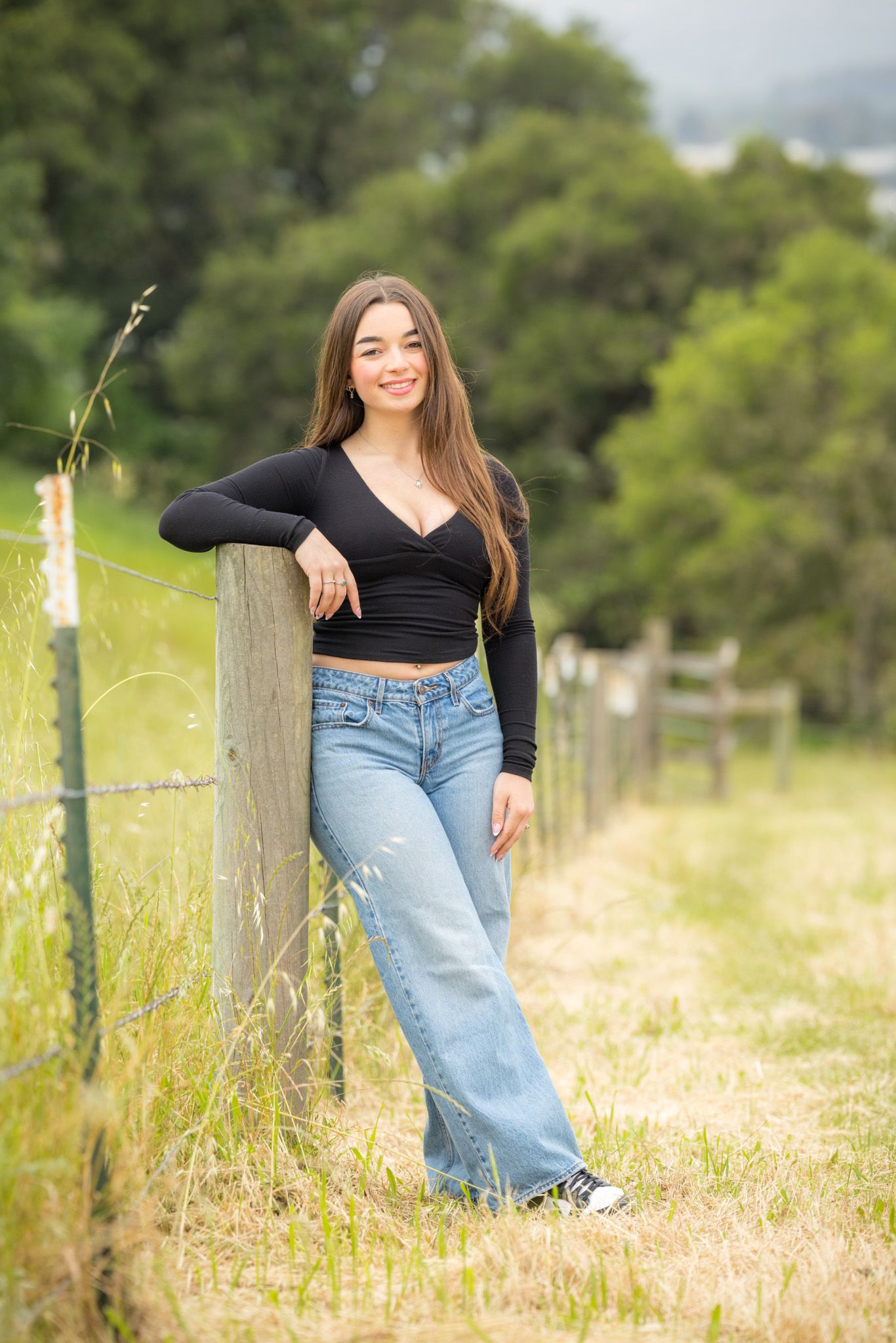 Young woman in black crop top and wide-leg jeans leaning against a rustic fence post in a green countryside setting.