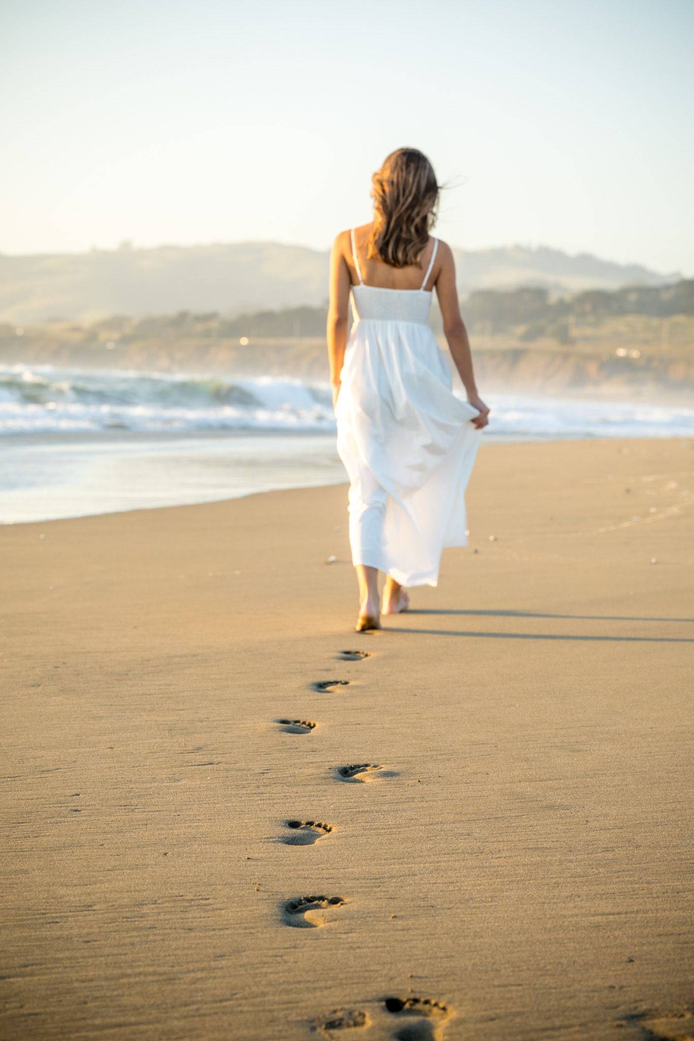 Woman in white dress walking barefoot on sandy beach at sunset, leaving footprints in the sand near ocean waves.