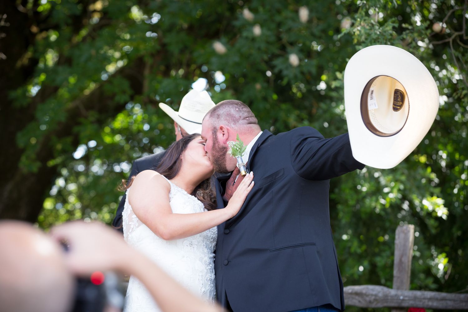 A couple shares a romantic kiss outdoors at a country wedding, with a cowboy hat visible in the sunlit garden setting.