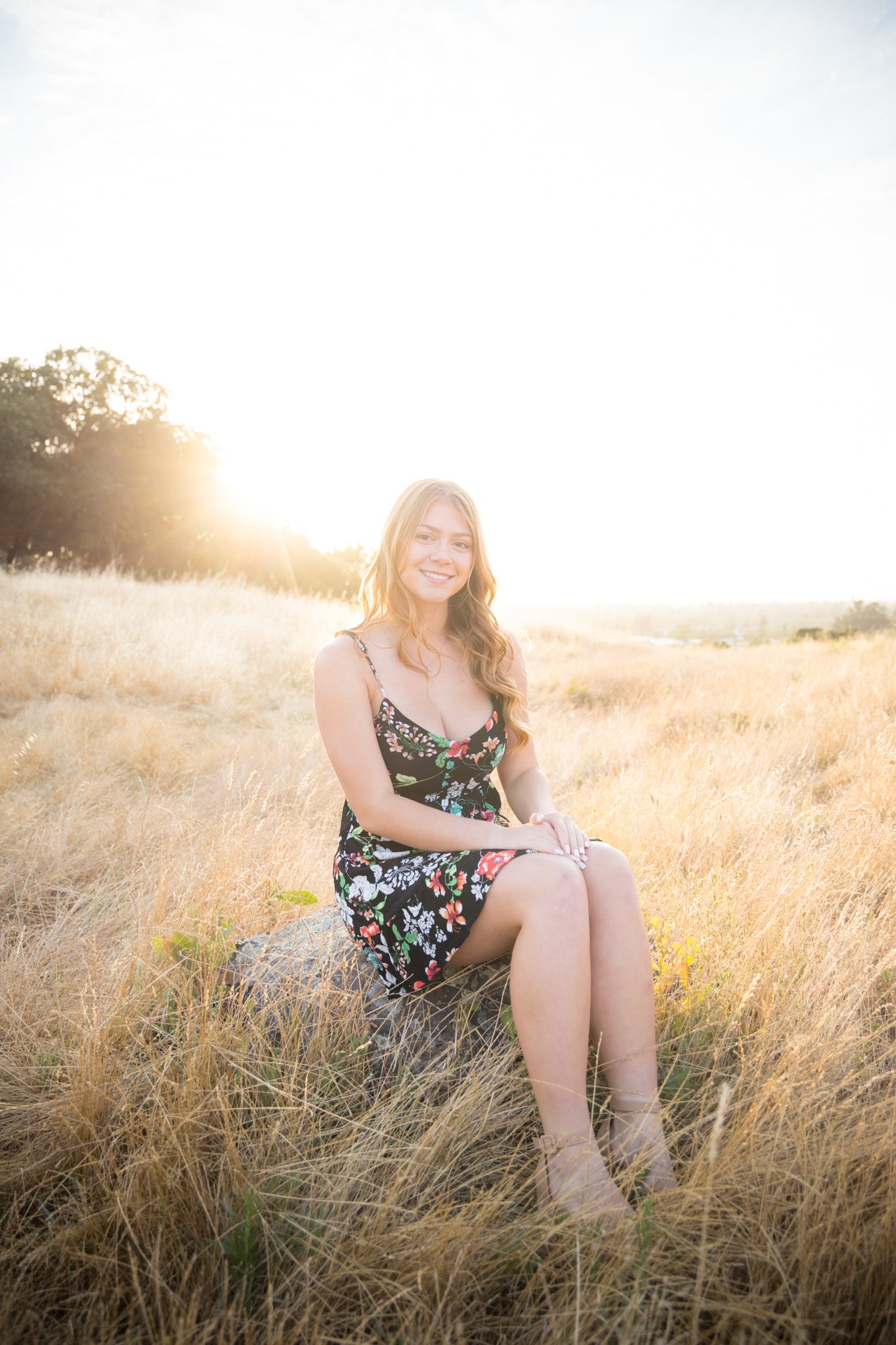 Golden hour portrait in a floral dress sitting in tall summer grass with warm backlighting.