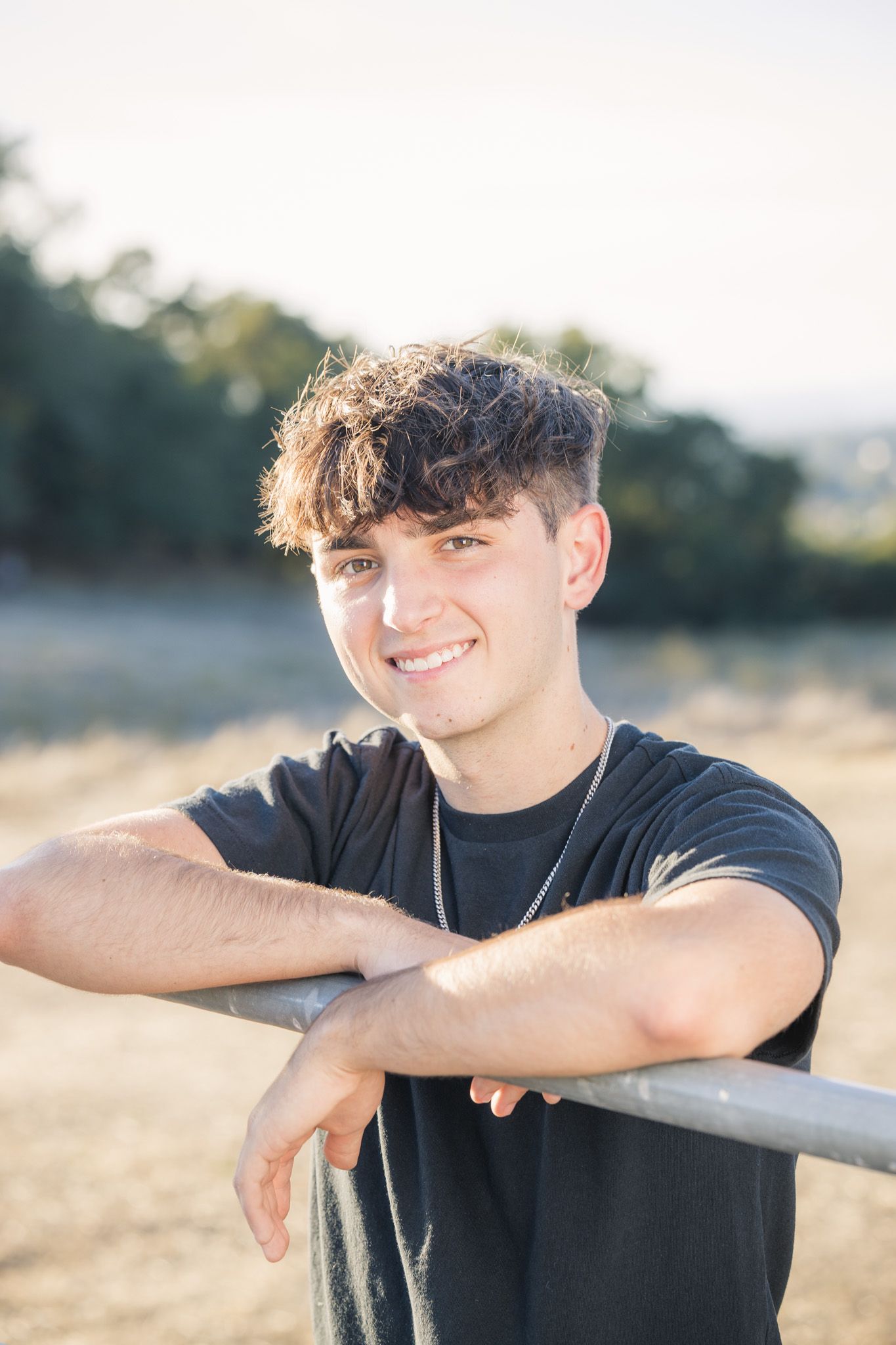 Young individual in black t-shirt leaning on railing during golden hour outdoor portrait session.