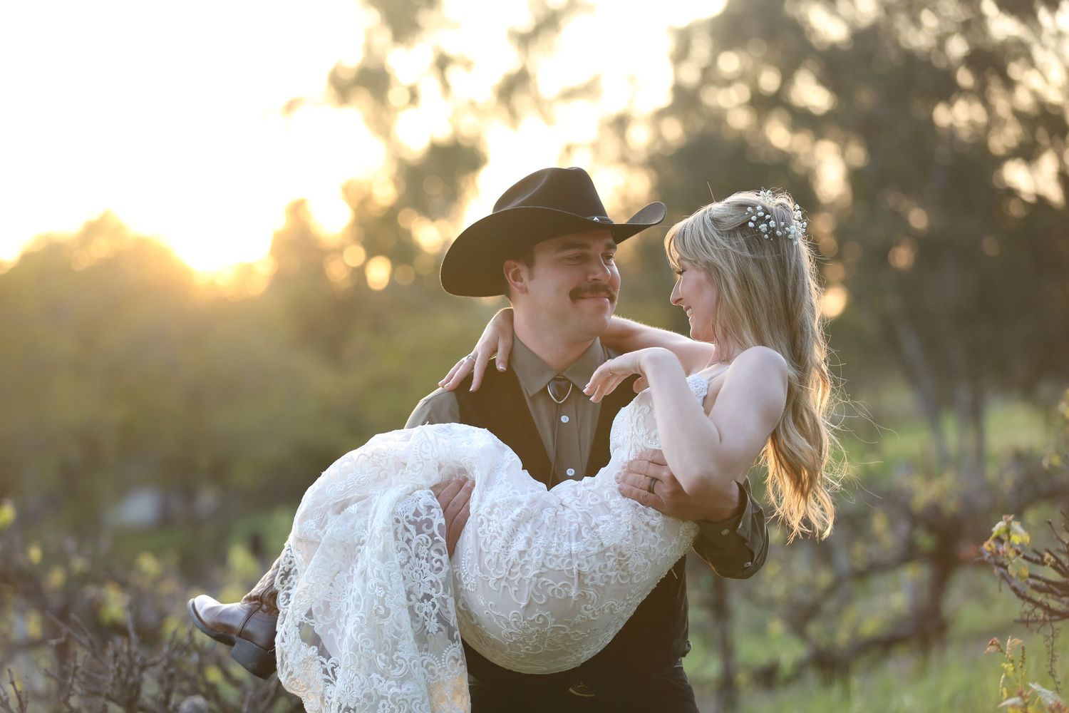 A romantic couple in western attire share an intimate moment at sunset, with the bride wearing a white lace dress.