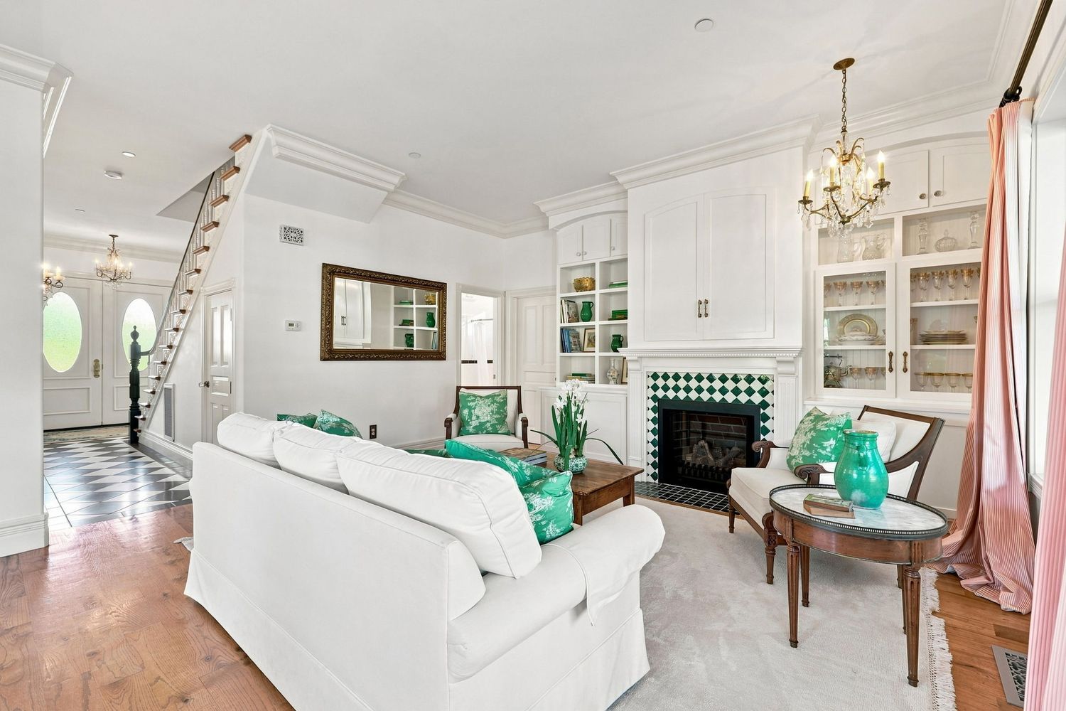 Bright white living room with green accents, built-in shelves, fireplace, chandelier, and staircase in elegant home.