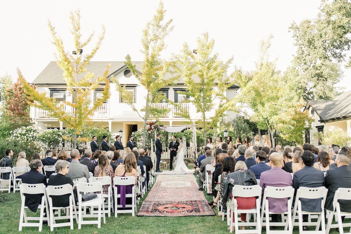 An outdoor wedding ceremony setup with white chairs and a red carpet aisle in front of a beautiful Victorian-style house.