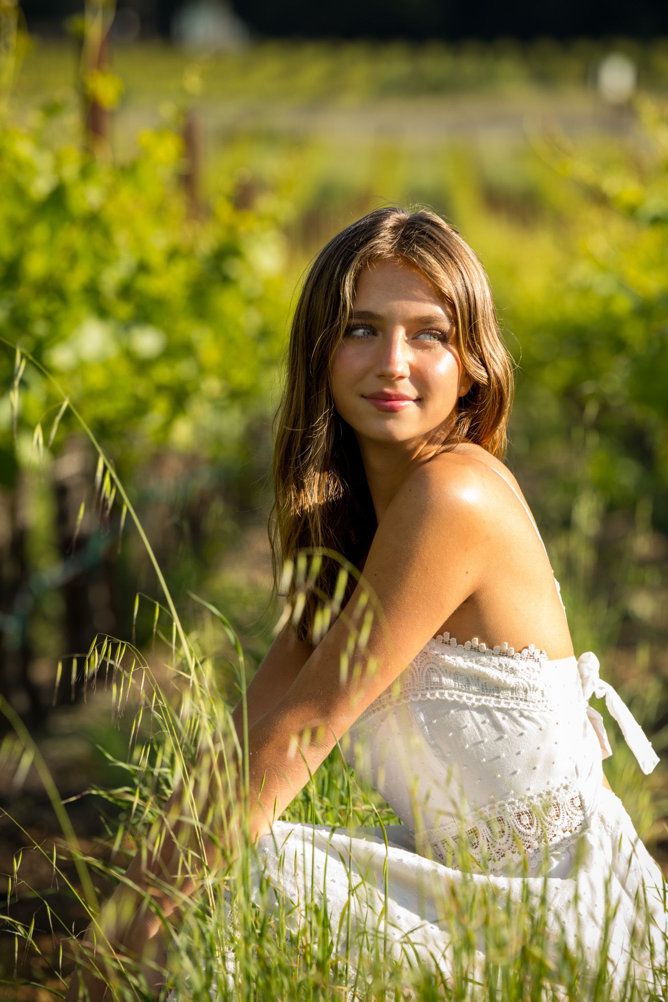 Young woman in white strapless dress sitting in lush green vineyard, long brown hair, golden sunlight.