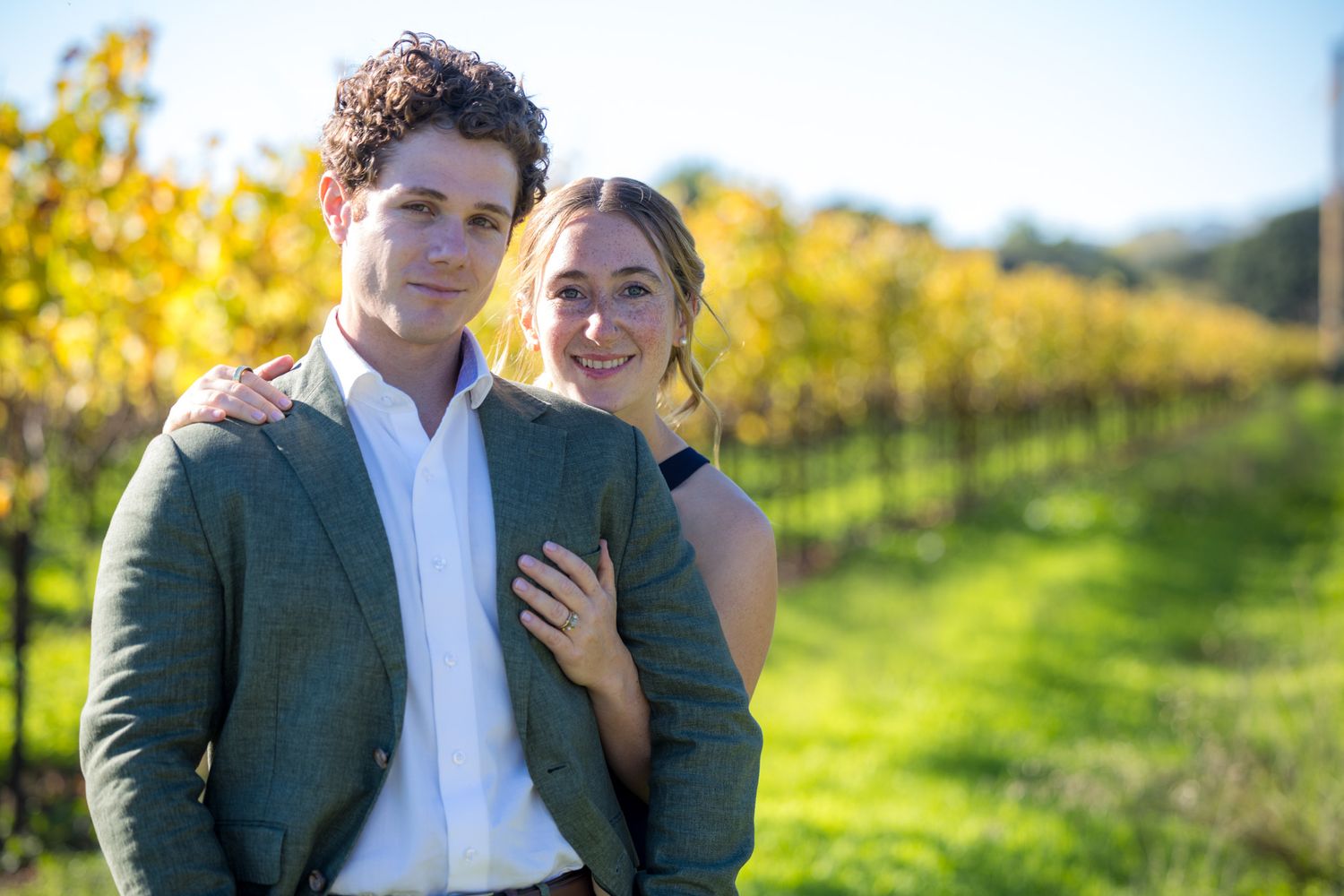 Couple posing together in a vineyard during autumn, wearing elegant attire with golden foliage in the background.