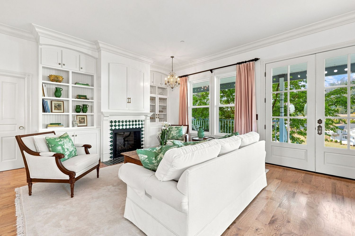 Elegant white living room with built-in shelves, green fireplace, white sofas, and French doors with pink curtains.