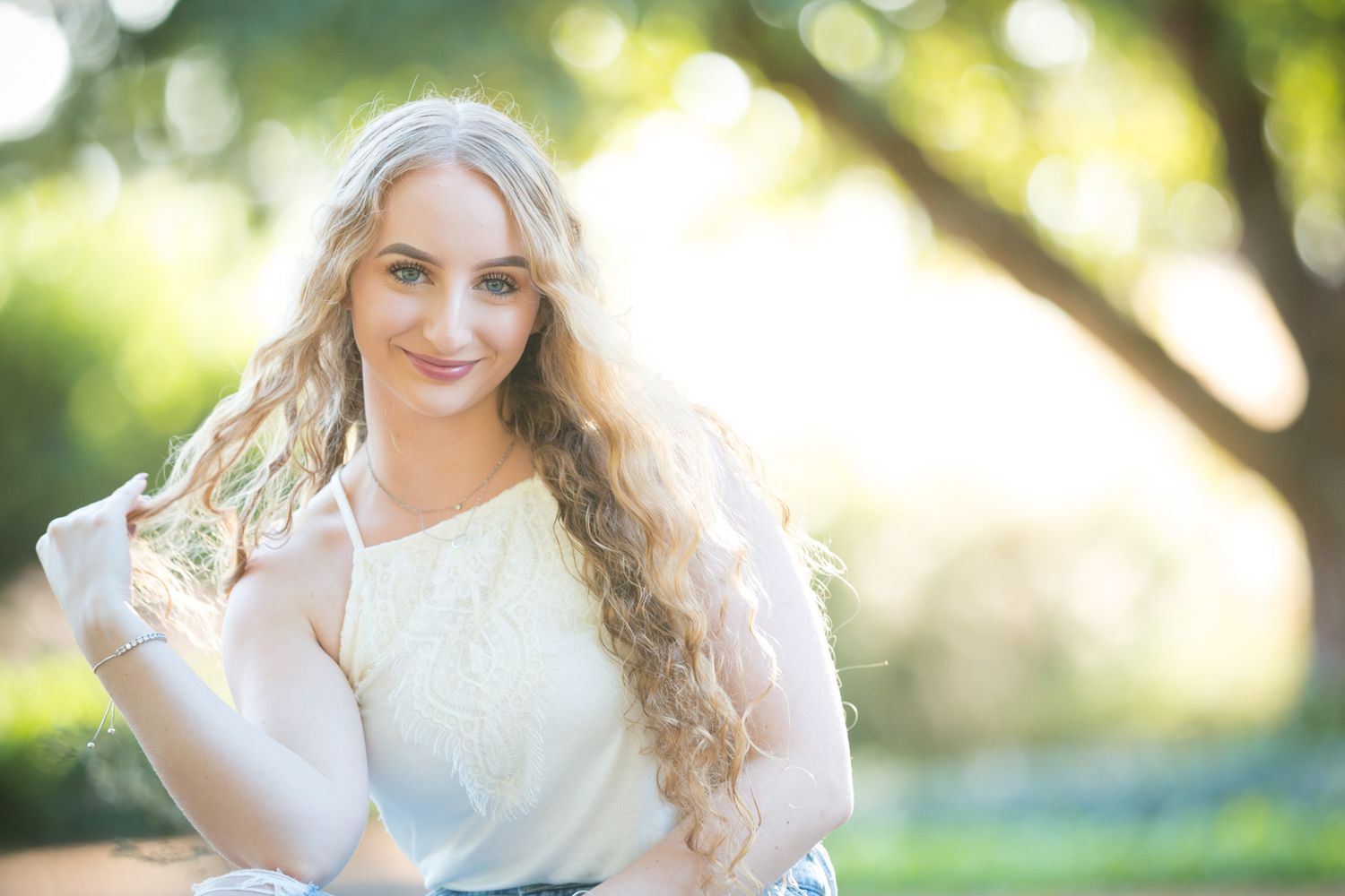 Outdoor portrait with soft lighting showing a woman in white dress with long blonde waves against a blurred natural backdrop.