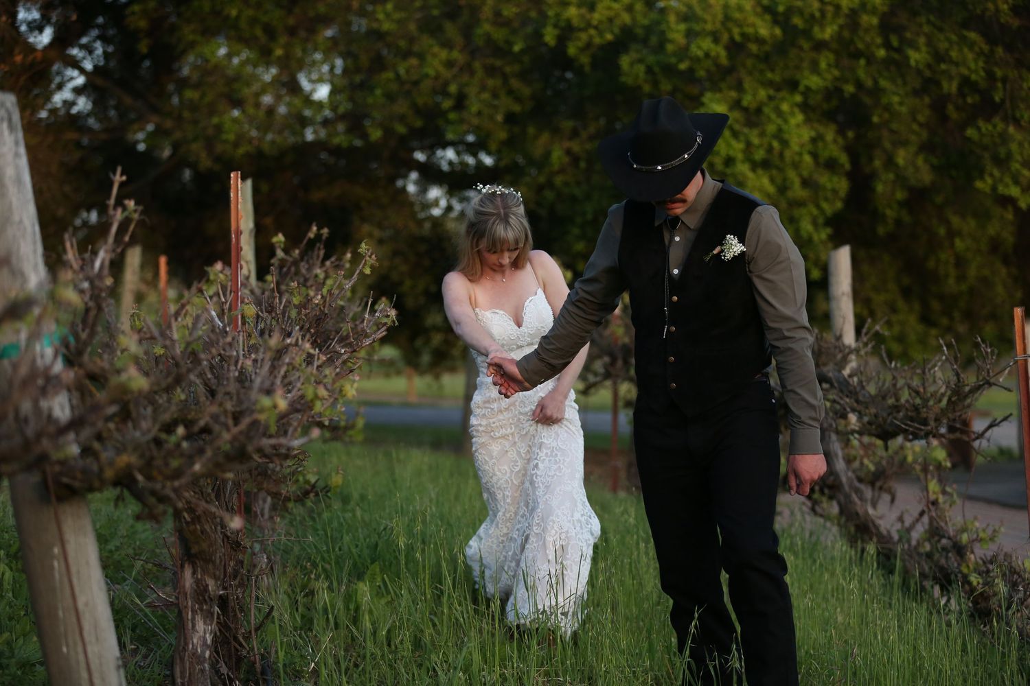 Couple walks together through a garden path at dusk, with bride in lace dress and groom in formal attire.