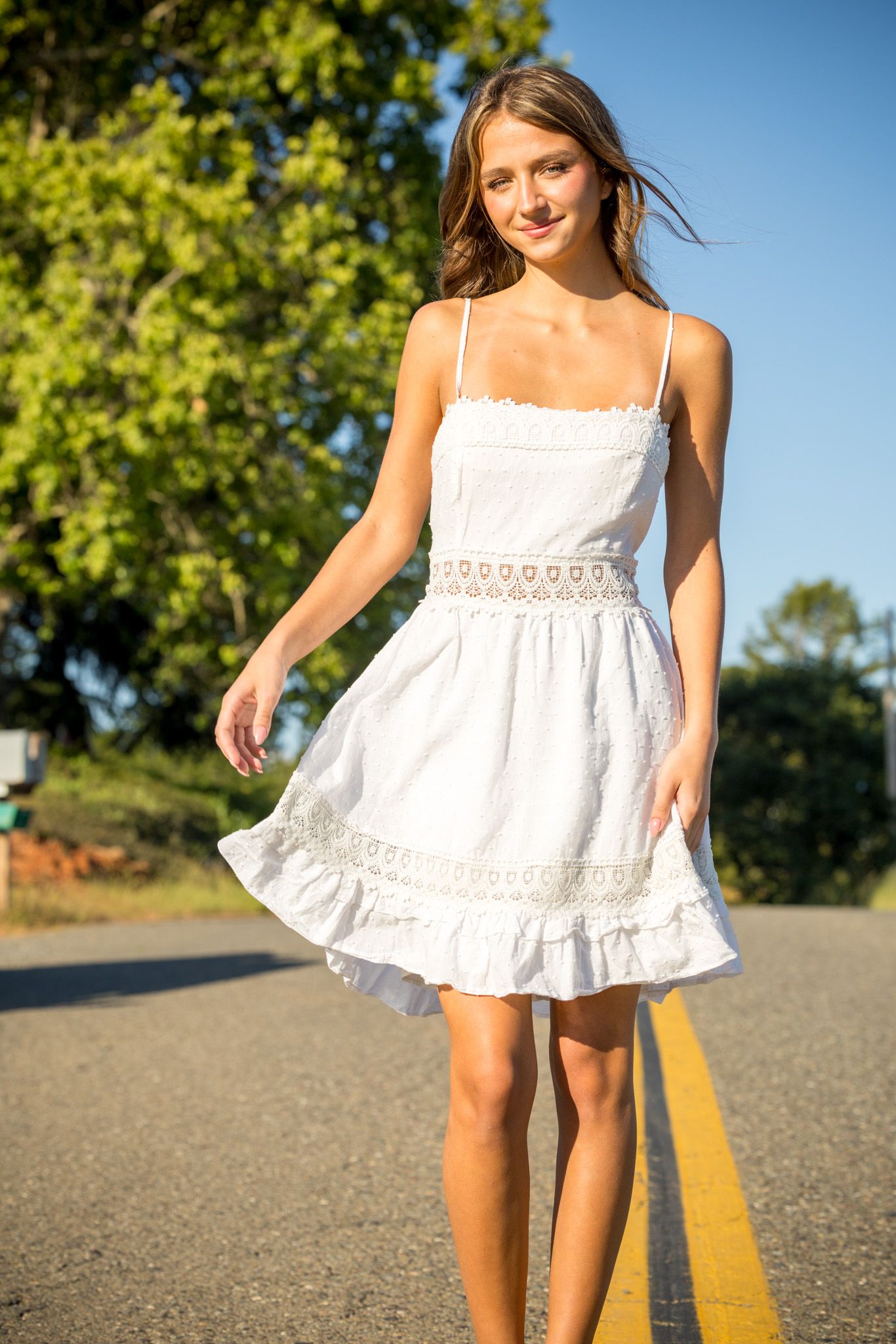 Young woman in white lace sundress walking on a sunny road surrounded by green trees and blue sky.