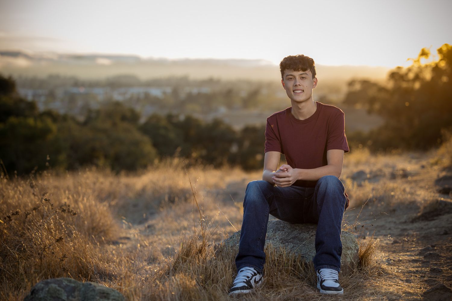 Senior Photos - Portrait of someone sitting on rocky terrain with sunset-lit hills in background.