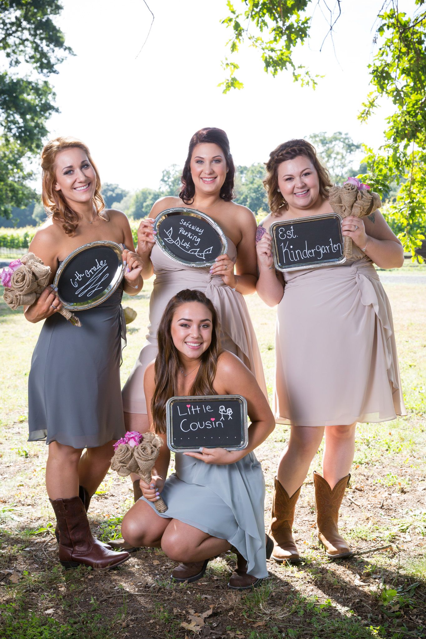 Bridesmaids in gray dresses and cowboy boots pose outdoors holding chalkboard signs in sunny garden setting.
