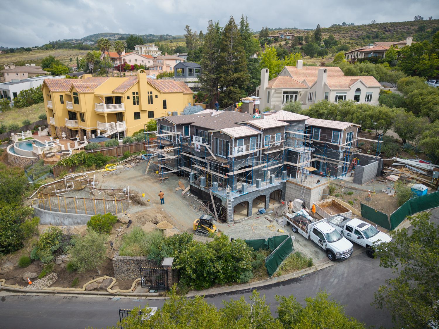 Aerial view of a large home under construction with scaffolding, surrounded by mature trees and neighboring houses.