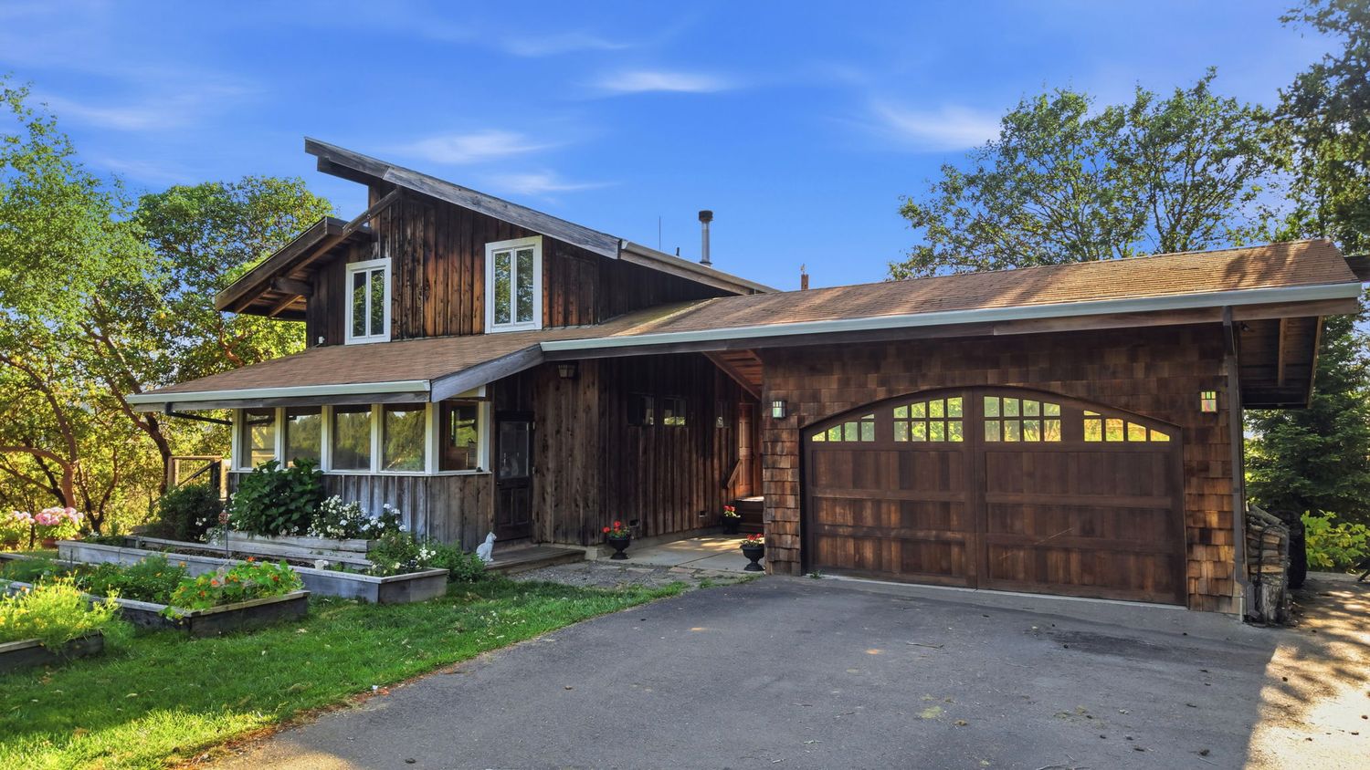 Rustic wood-sided home with attached two-car garage, covered porch, and mature trees on a paved driveway.