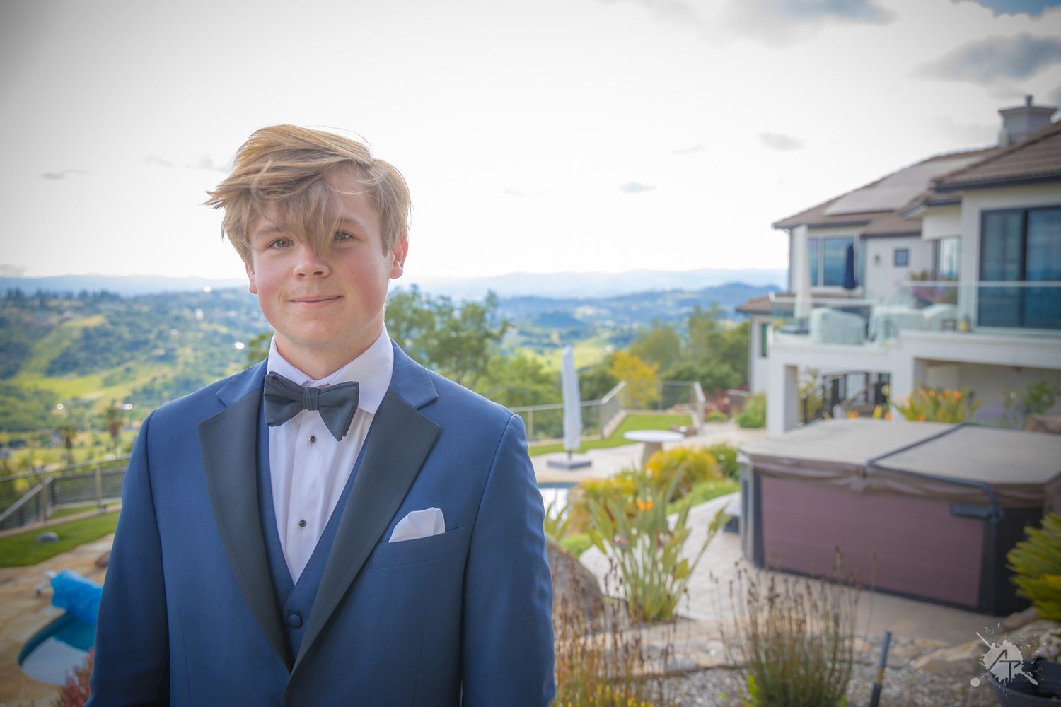 Person in classic black tuxedo and bow tie poses at scenic overlook.