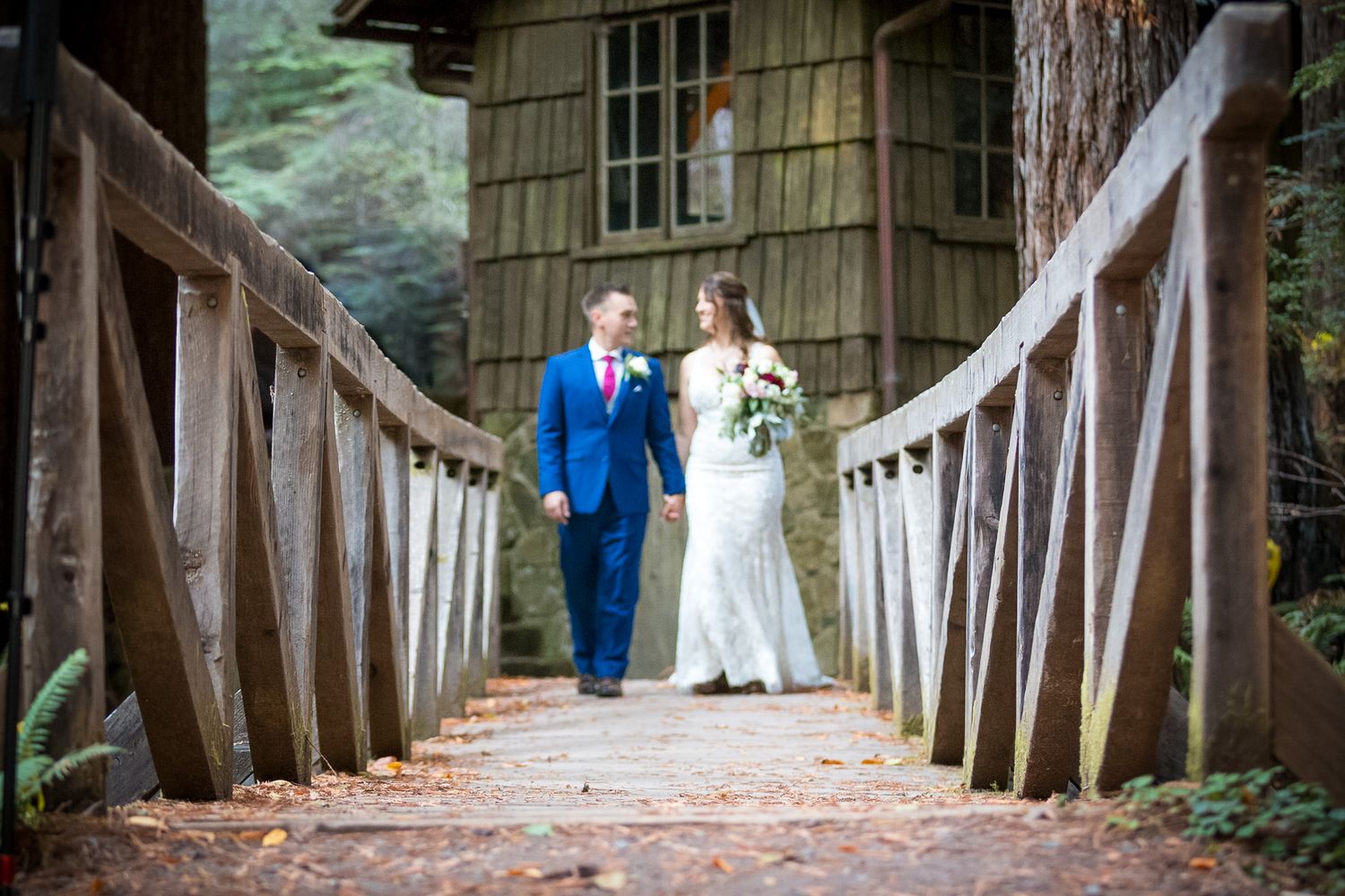 Wedding couple stands on a wooden bridge with rustic cabin in background surrounded by forest.