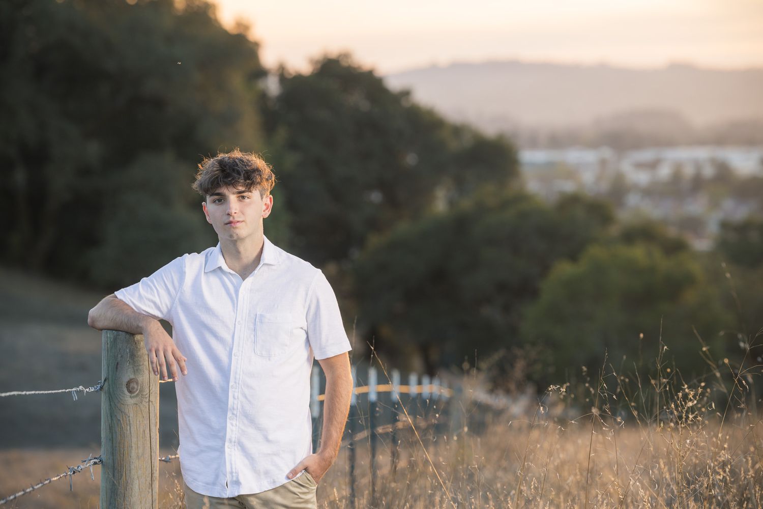 Person in white shirt leaning on wooden fence post during sunset with hills in background.