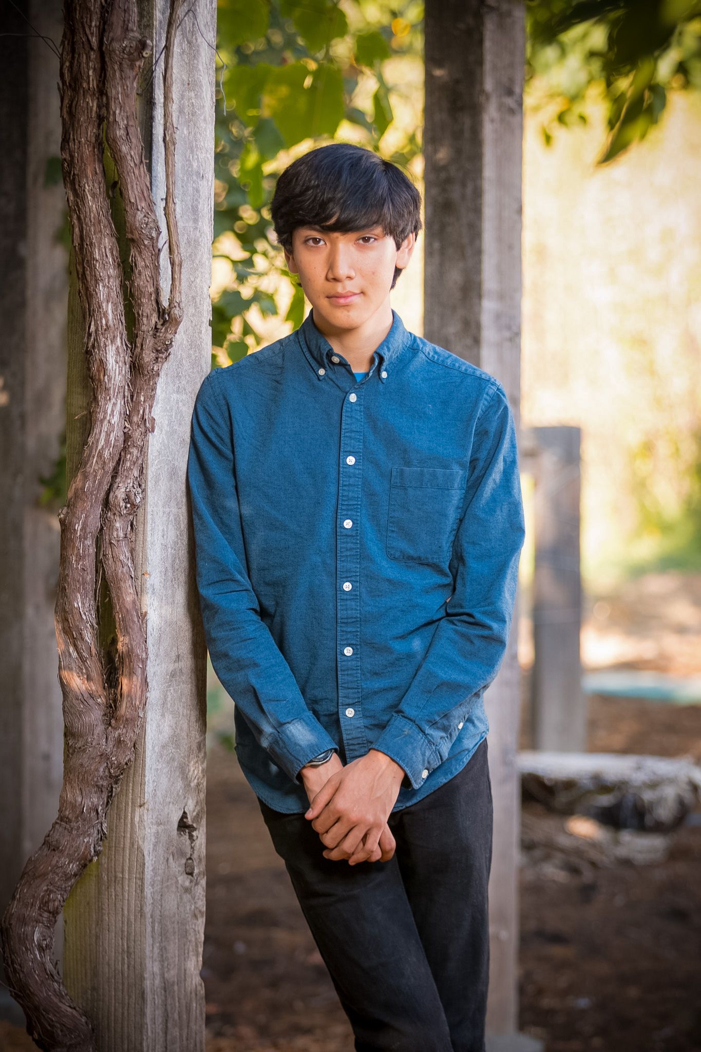 Portrait of someone in a blue button-down shirt standing against a wooden pillar in natural lighting.