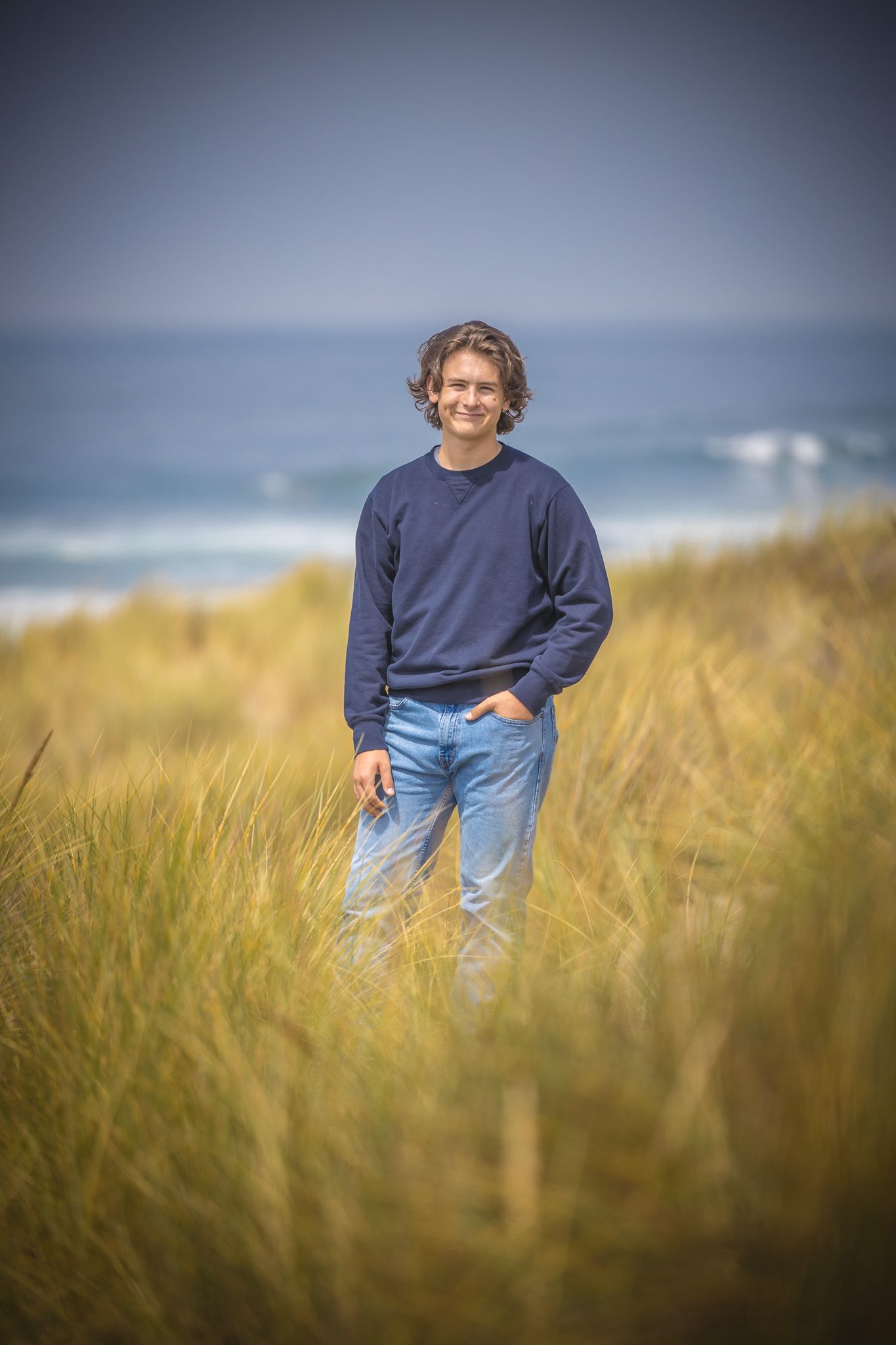 Person standing in tall beach grass wearing navy sweater with ocean and cloudy sky in background.