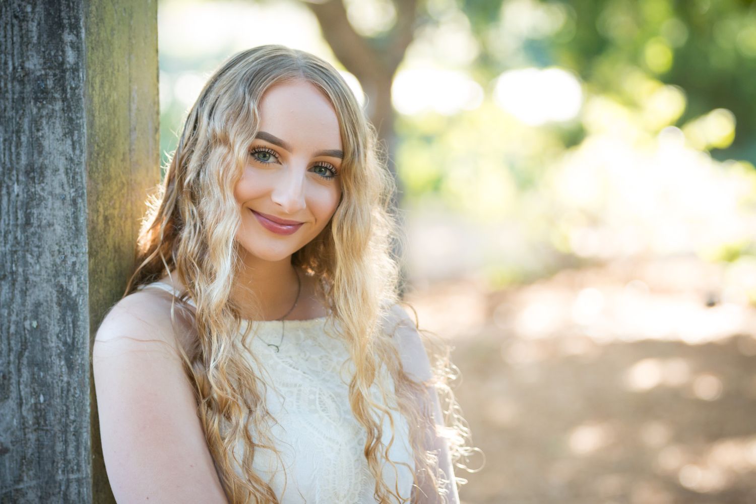 A portrait of someone with long wavy blonde hair and a warm smile wearing a white lace dress outdoors in soft lighting.