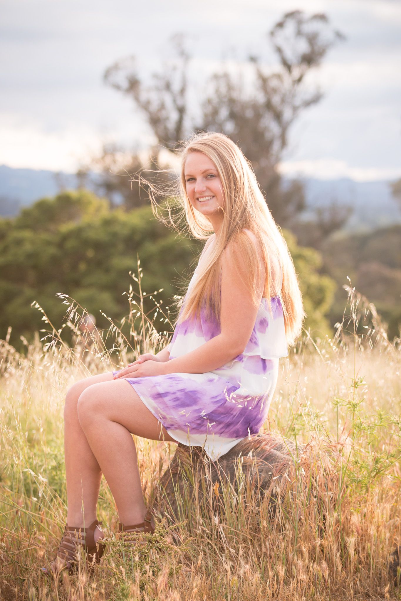 Person in purple tie-dye dress sits on hillside during golden hour with mountains and dry grass in background.