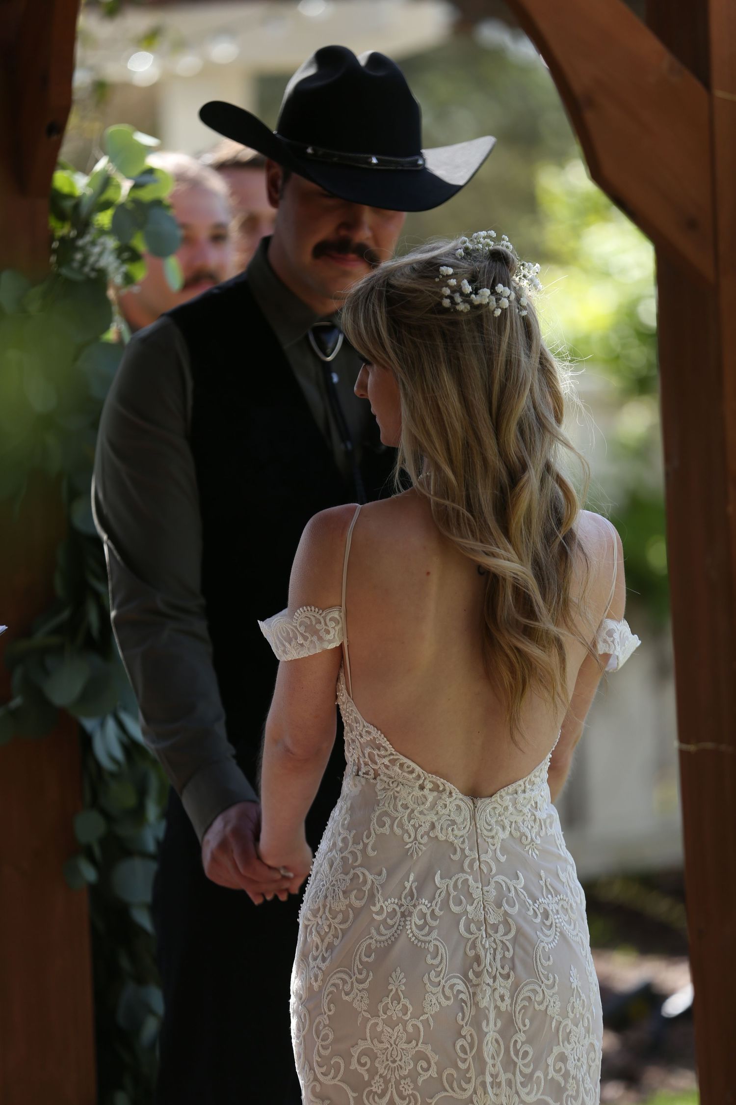 A couple shares a moment during their wedding ceremony under a wooden arch with lace dress details and western attire.
