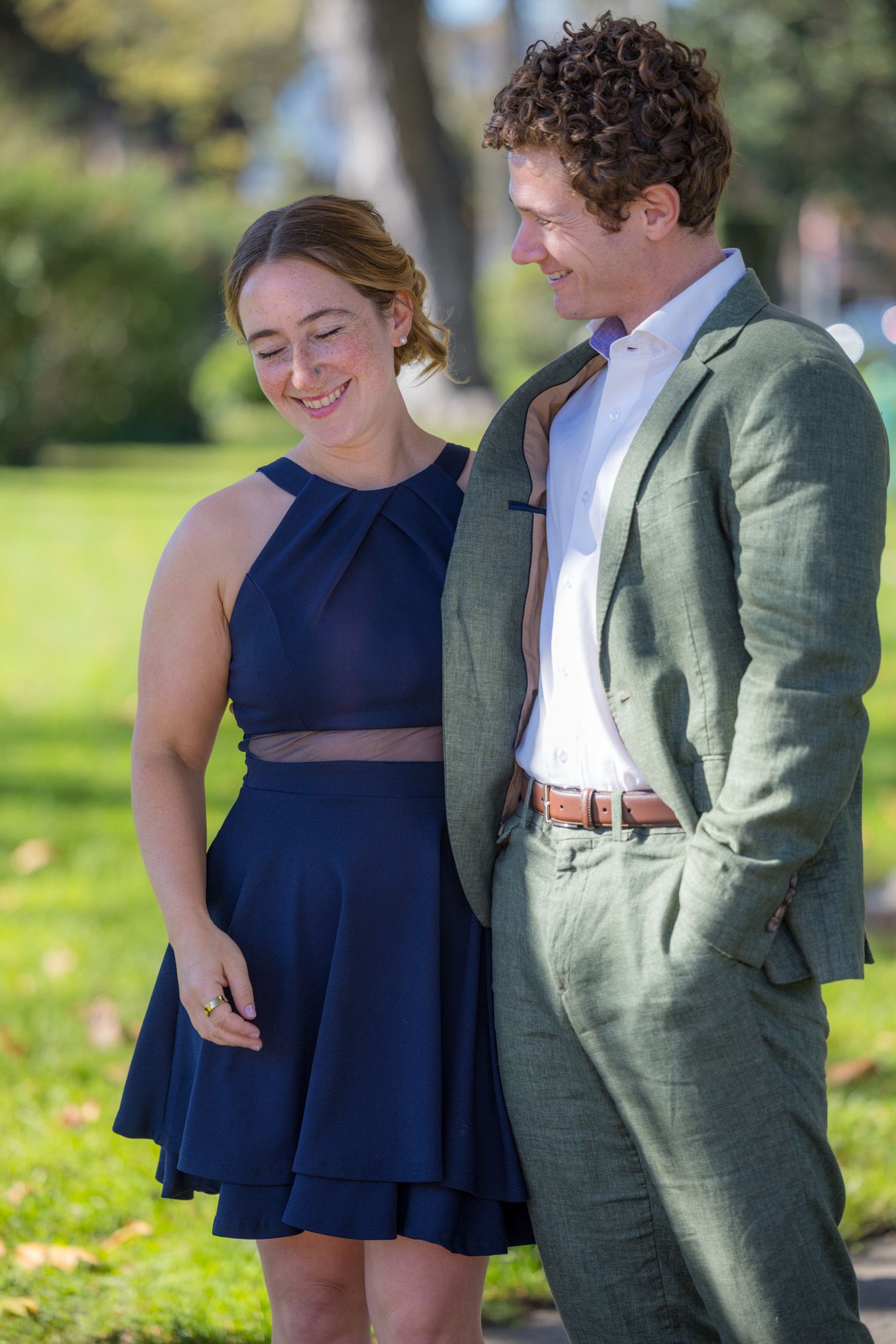 A smiling couple in formal attire poses outdoors, woman in navy dress and man in green suit, in a sunny park.