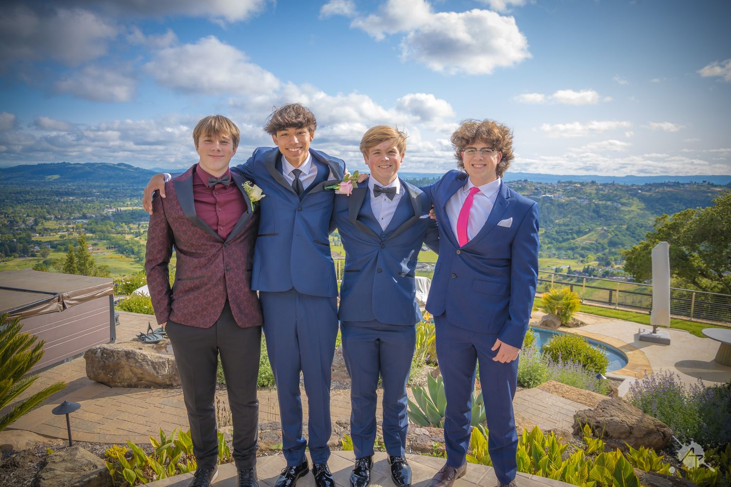 Four friends in formal attire pose together against a scenic mountain backdrop on a sunny day.