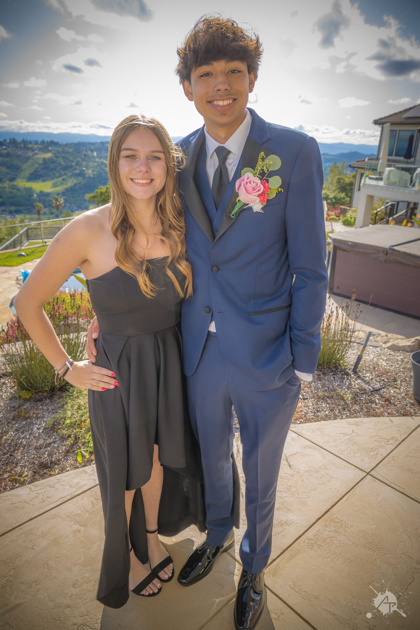 A couple dressed for a formal event poses on an outdoor patio with mountain views.