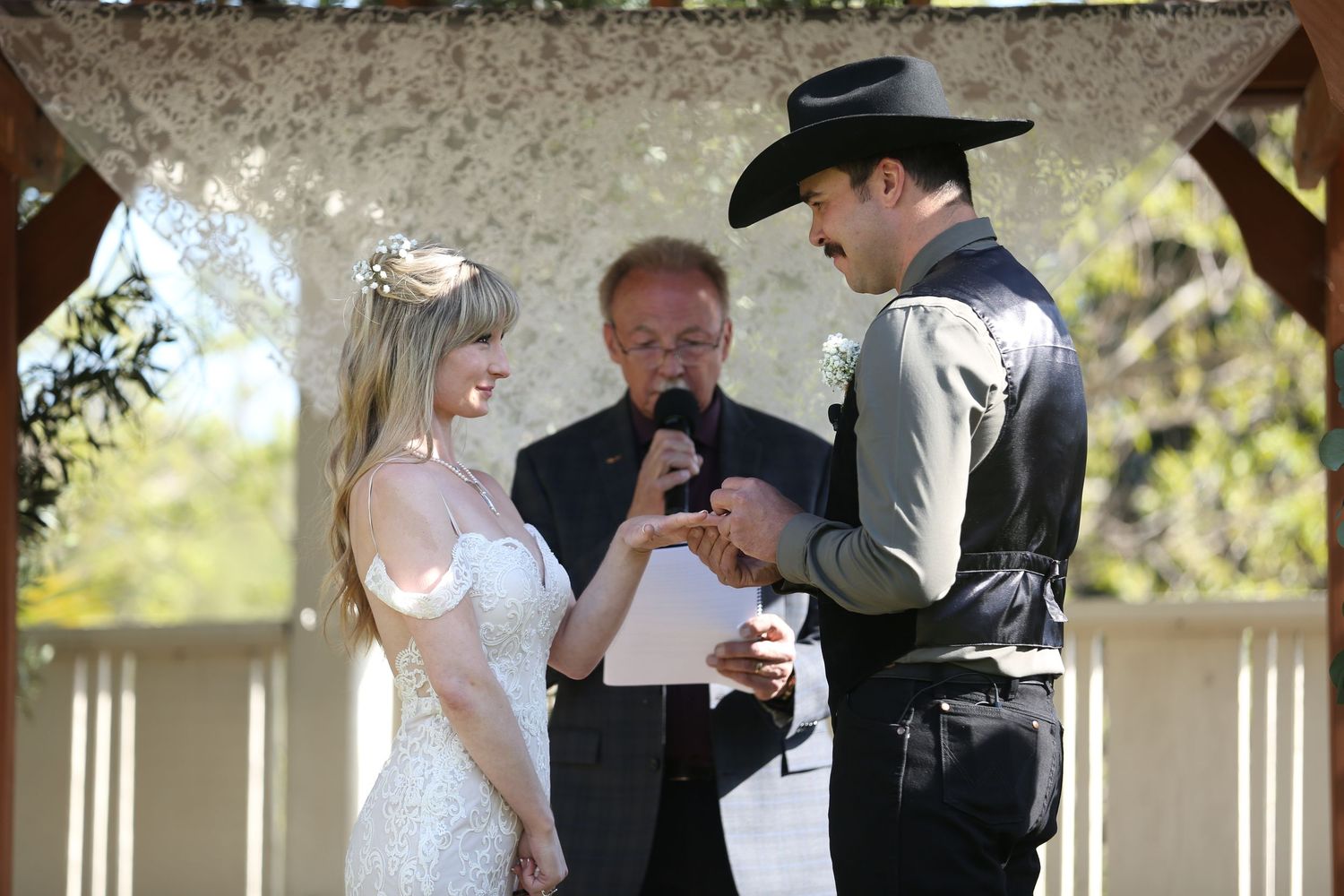 An outdoor wedding ceremony takes place under a wooden gazebo on a bright, sunny day.
