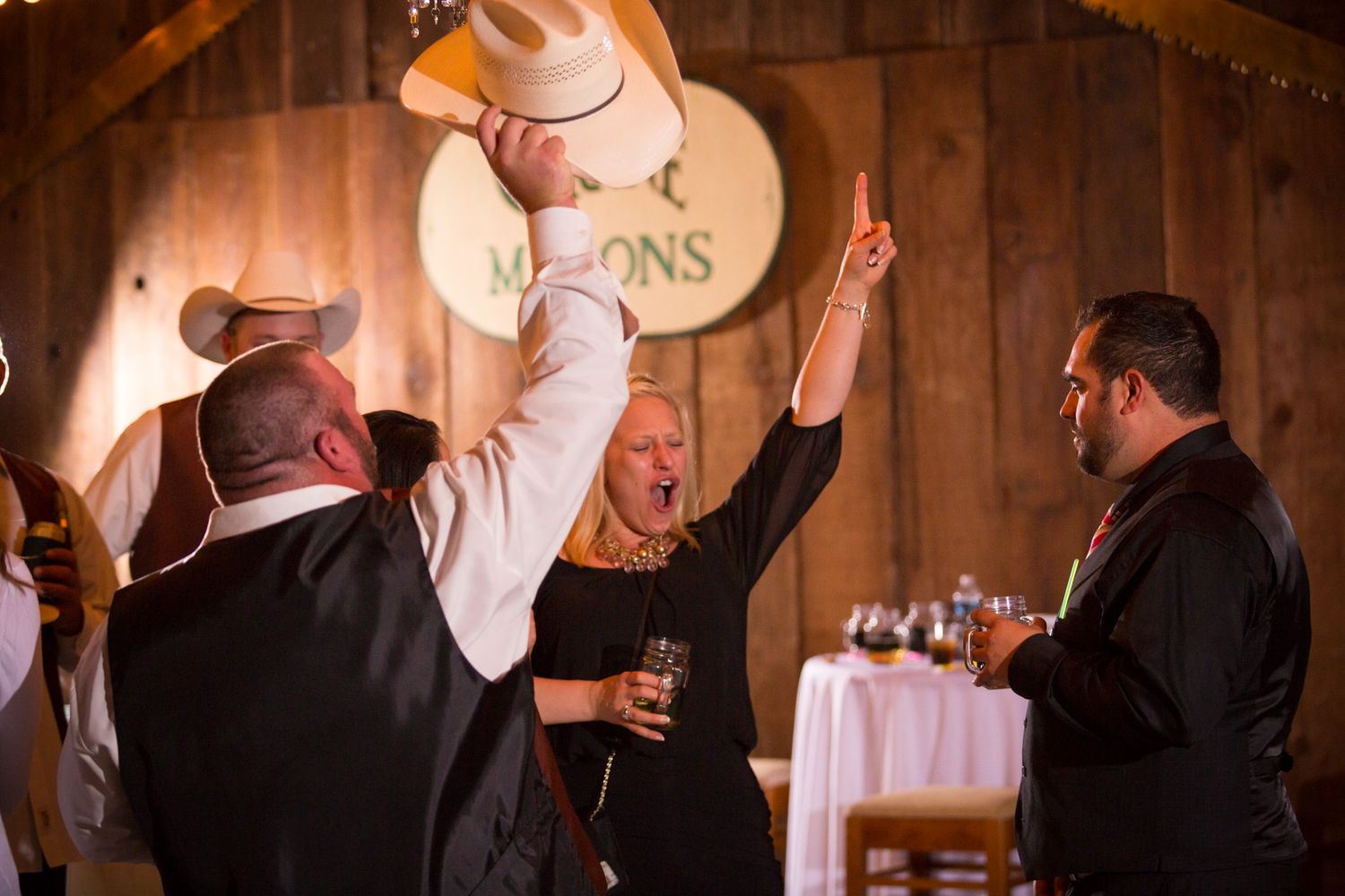 Wedding guests celebrate and dance at a rustic barn reception with wooden walls and ambient lighting.