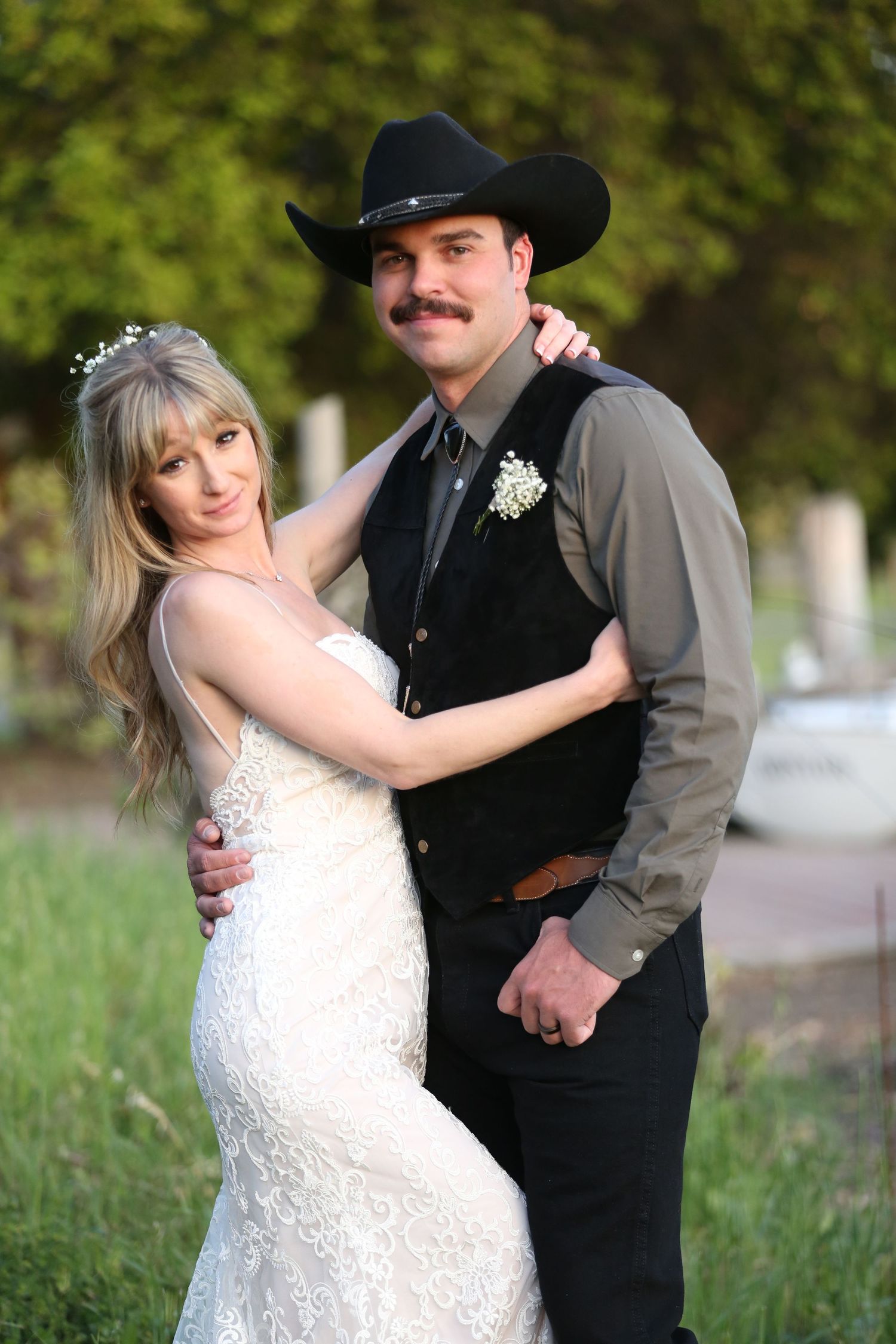 Romantic wedding couple embraces outdoors, bride in lace gown and groom in black cowboy hat and western attire.