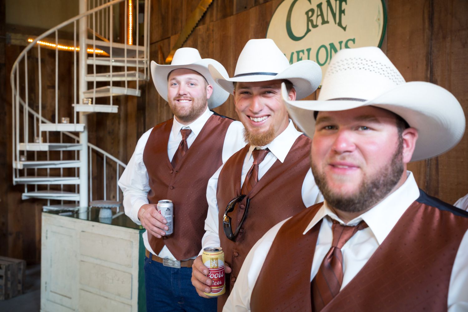 Three groomsmen in matching brown vests and white cowboy hats smile together outside a rustic wooden venue.
